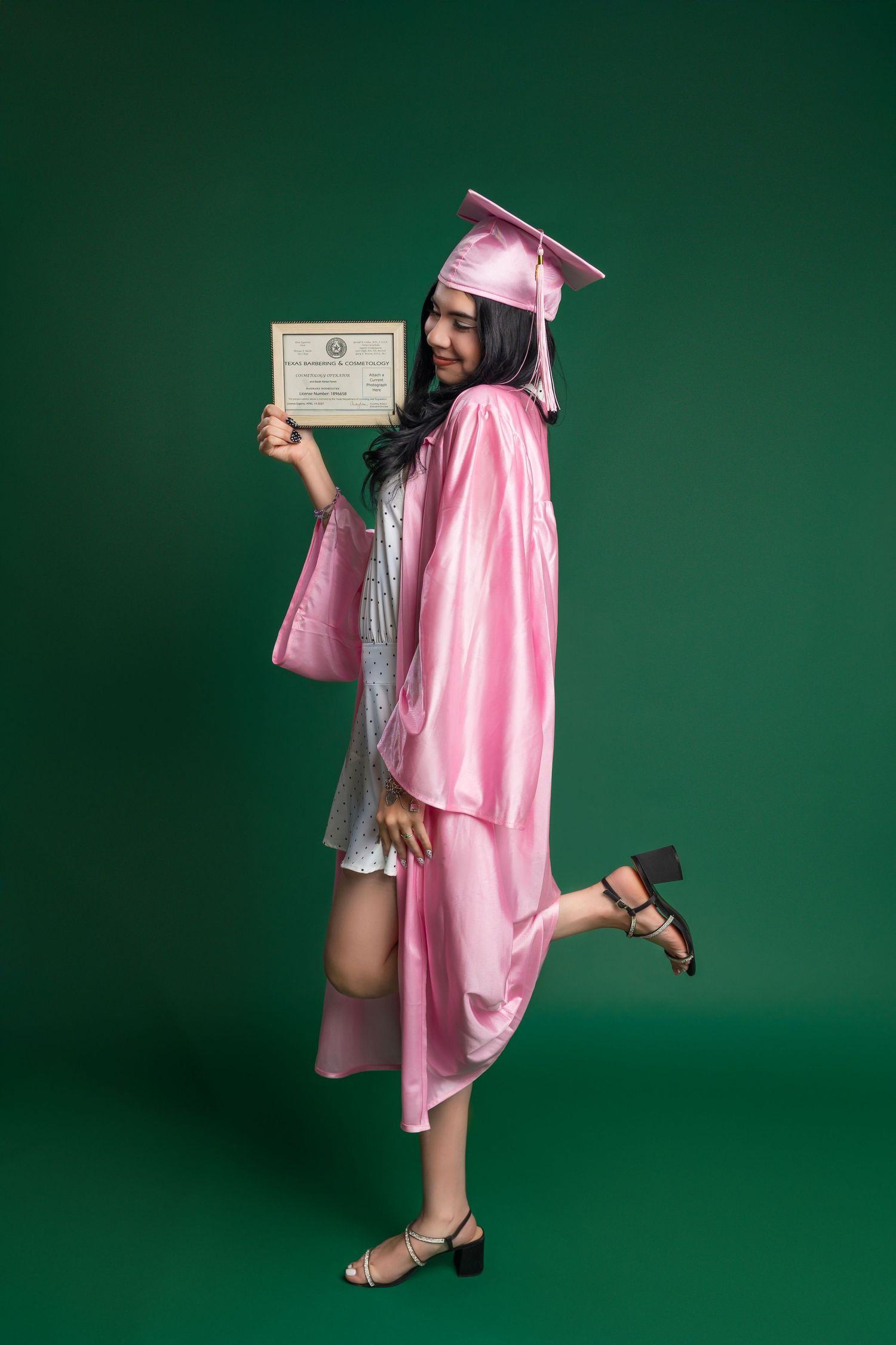 Graduate in pink cap and gown poses playfully while holding up diploma certificate against green background.