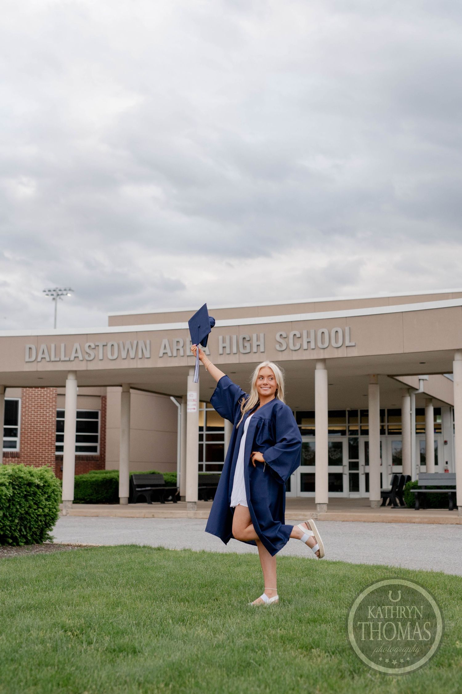 A graduation photo at Dallastown Area High School entrance with cap toss and blue graduation gown against cloudy sky.