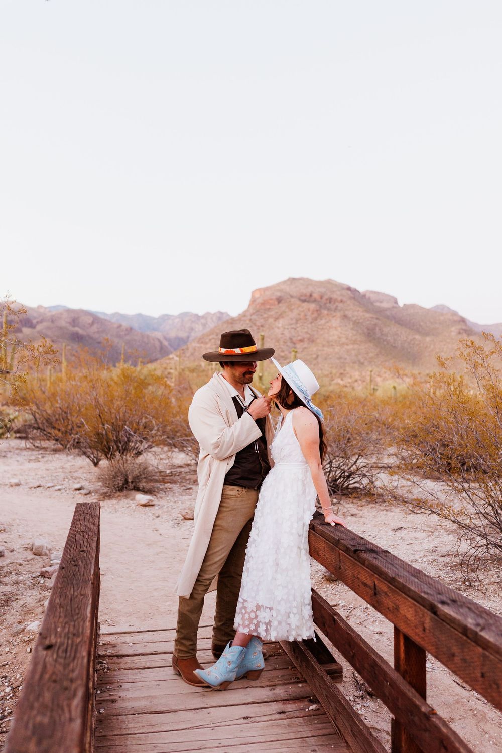 A couple embraces on a wooden walkway in the desert with mountains and brush in the background at sunset.
