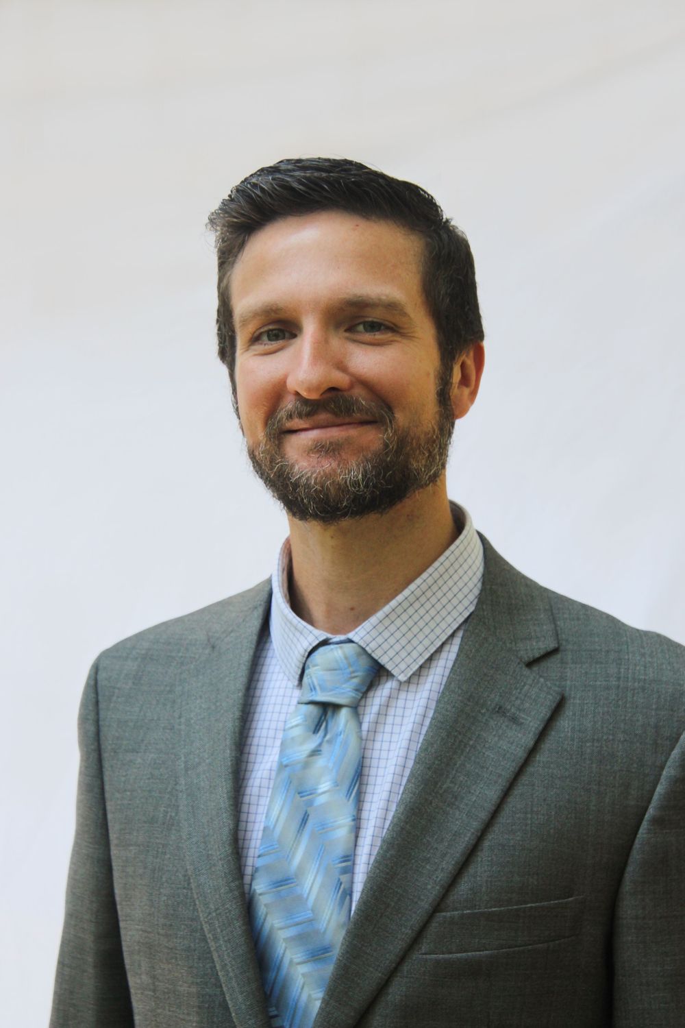 Professional headshot of a person wearing a grey suit jacket, checkered dress shirt, and light blue patterned tie.