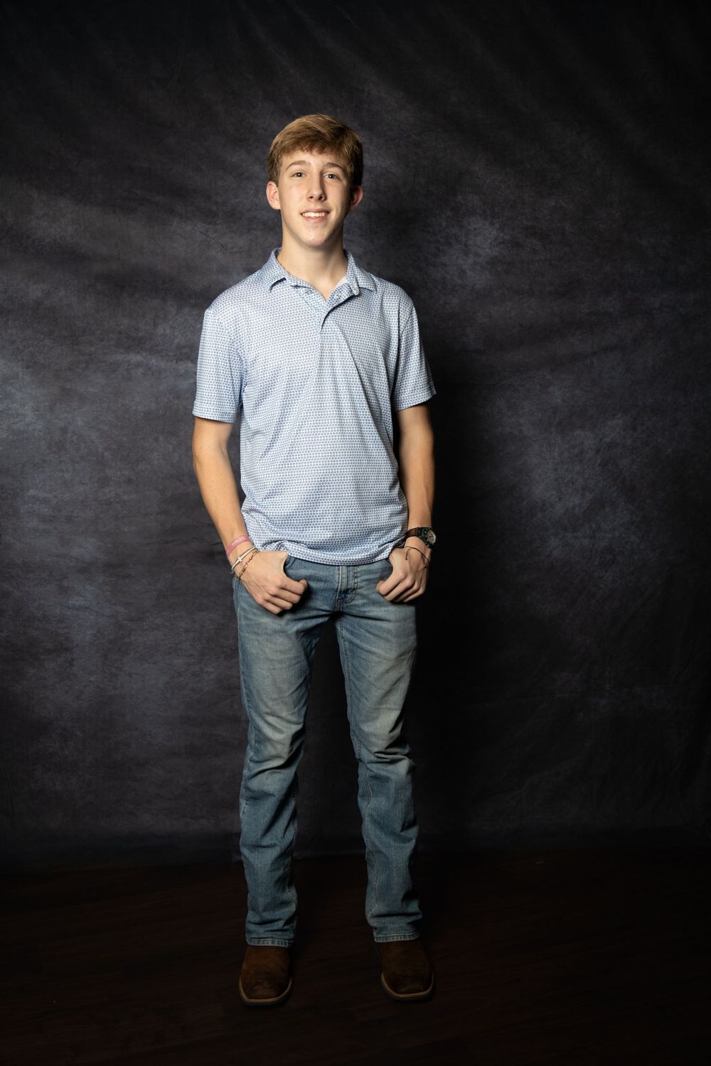 A teenage boy in a light blue polo shirt and jeans stands smiling with hands in pockets against a dark studio backdrop.