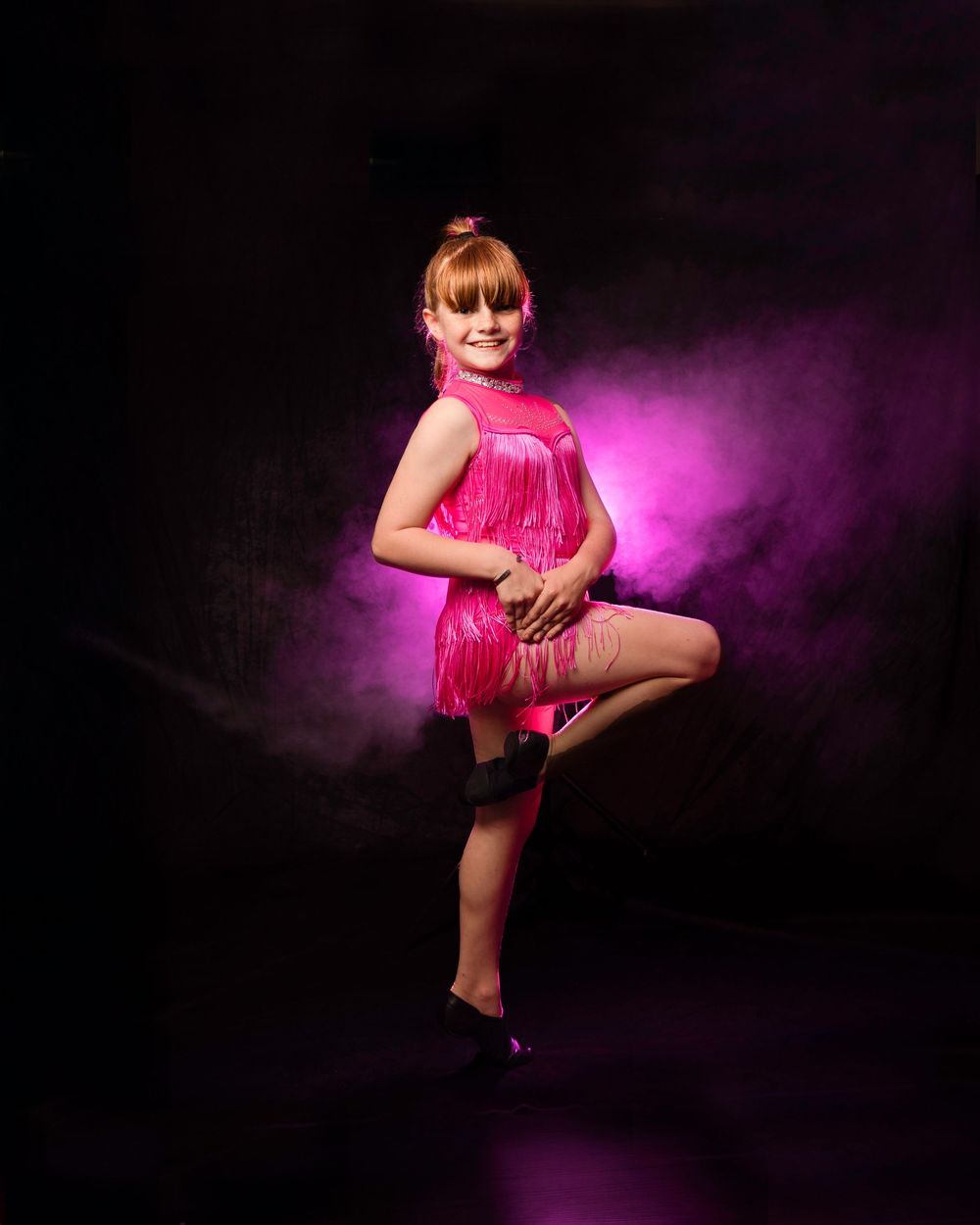 Young girl in pink fringe dance costume poses gracefully on one leg against dramatic purple-lit dark background.