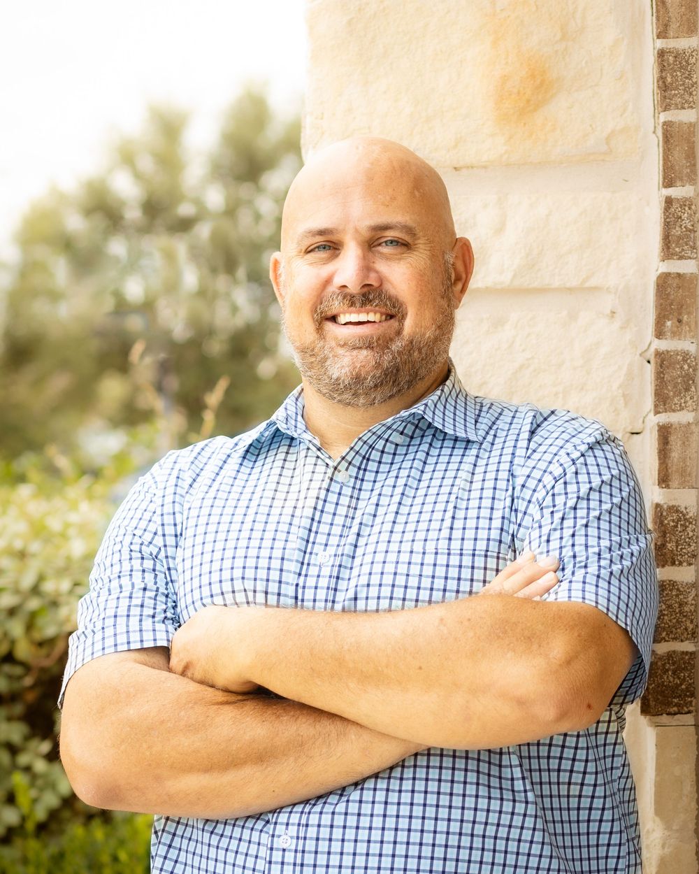 Man wearing a blue checkered shirt with crossed arms standing against a beige wall in outdoor lighting.