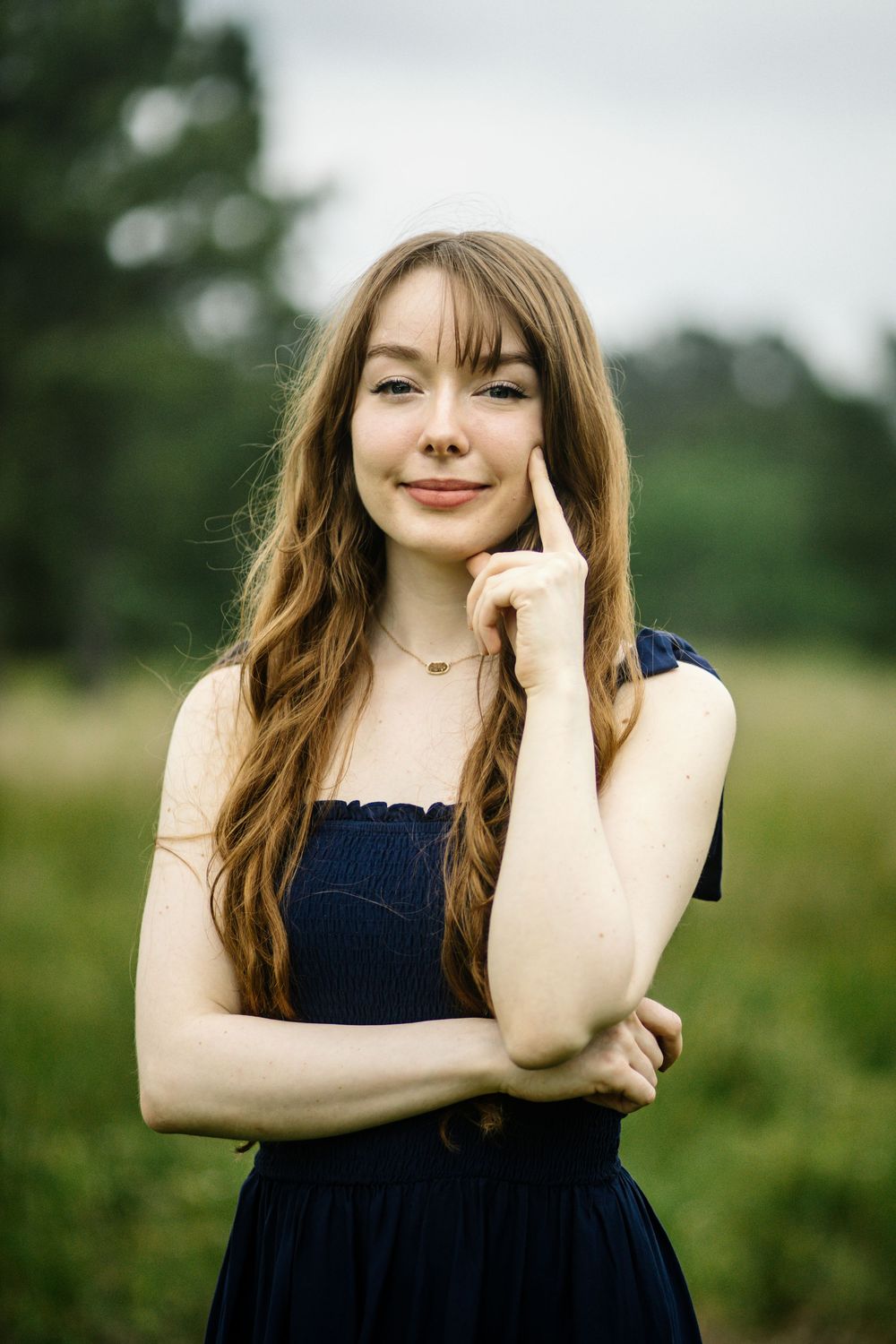 A young woman in a navy blue dress poses thoughtfully outdoors against a green natural background.
