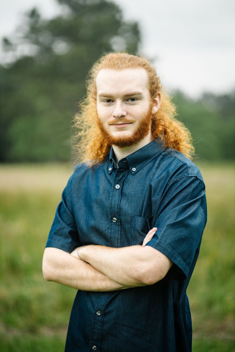 Person with long red hair and beard wearing a navy button-down shirt stands confidently with arms crossed in a grassy field.