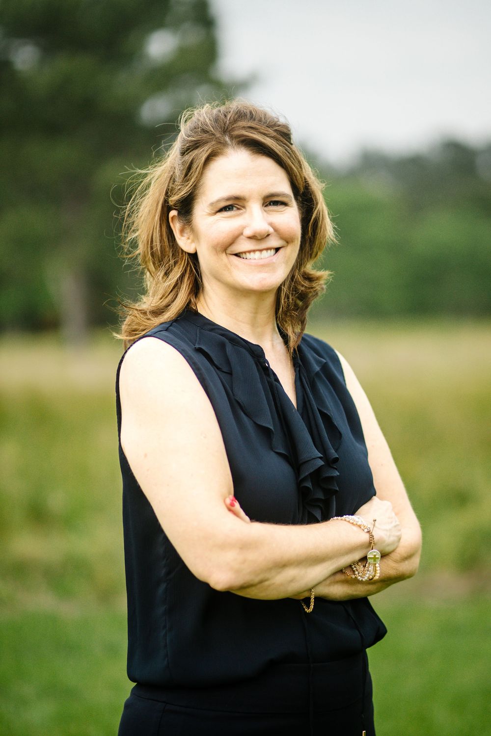 Professional portrait of someone in a black sleeveless blouse standing outdoors with arms crossed and natural backdrop.
