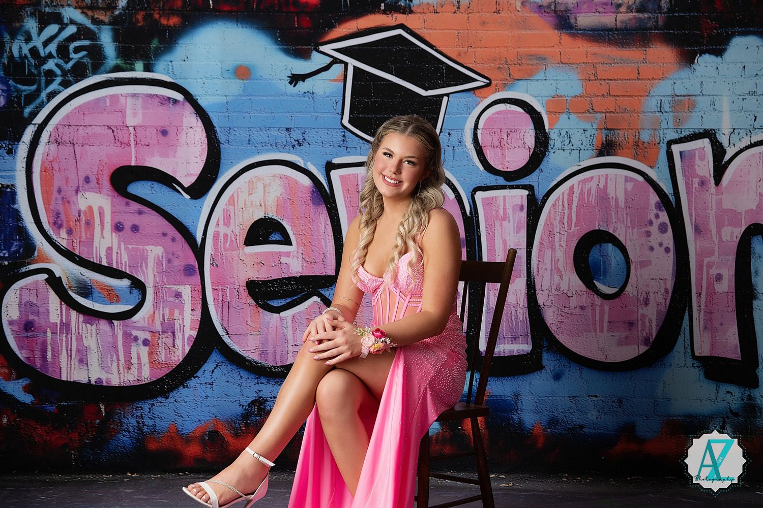 Graduate in cap and pink dress poses against colorful graffiti wall with 'Senior' text backdrop.