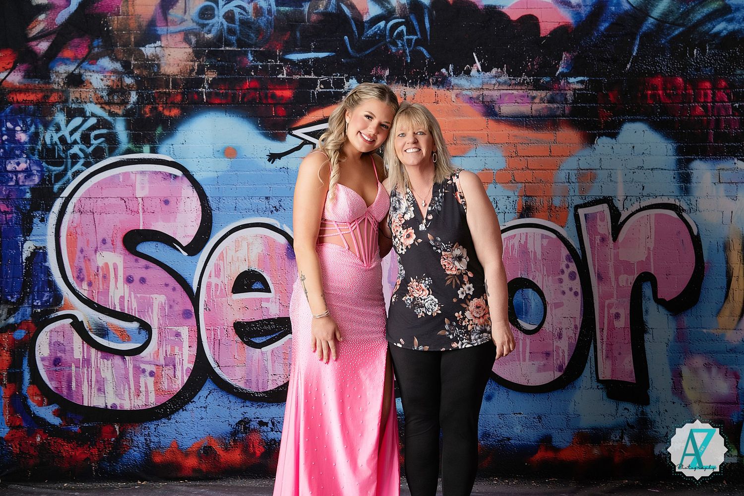 Two people stand posing in front of a colorful graffiti wall with pink text spelling 'Senior' in the background.
