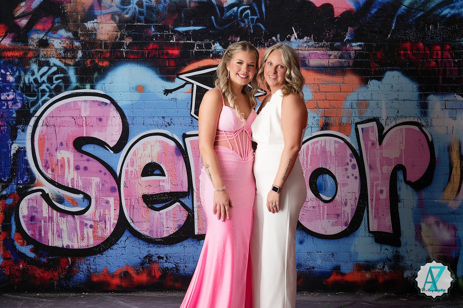Two people in formal dresses pose together in front of a colorful graffiti wall with pink text.