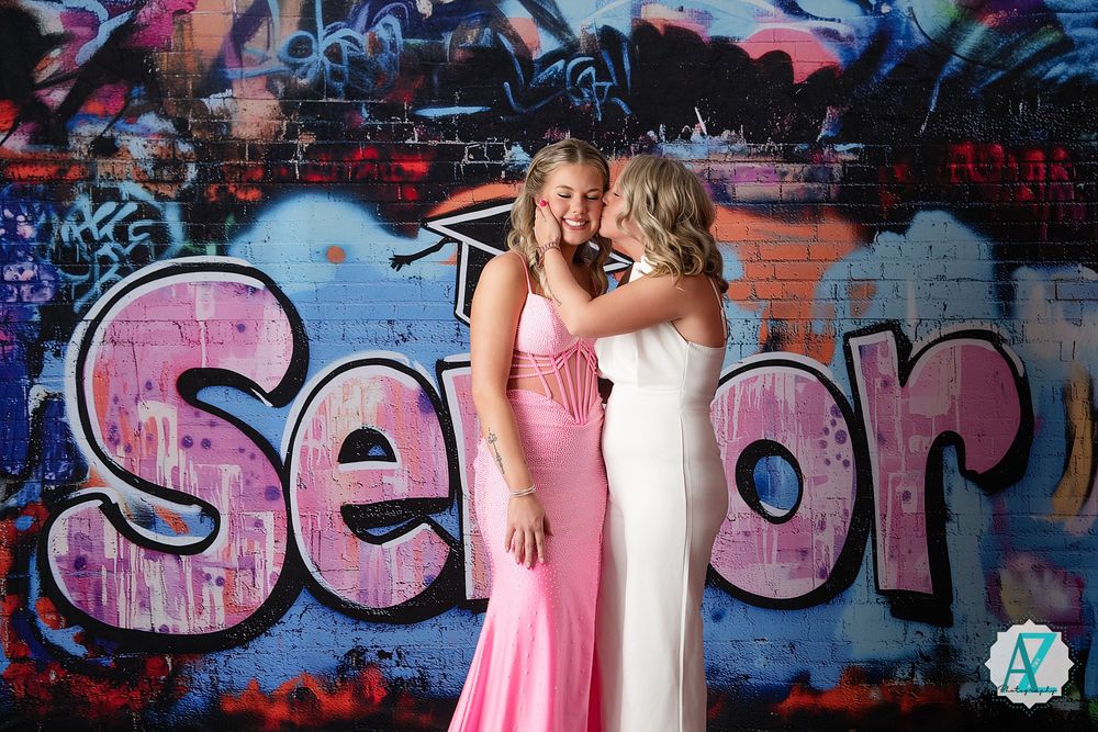 Two people in elegant dresses pose together against a colorful graffiti wall with pink spray paint text.