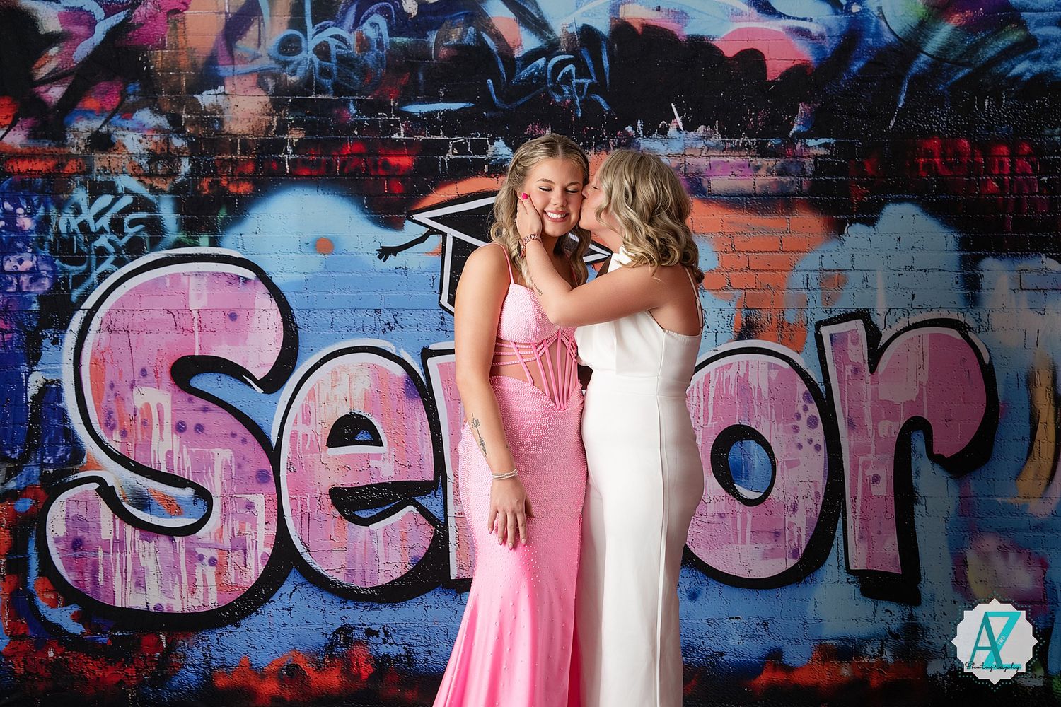 Two people in elegant dresses pose together against a colorful graffiti wall with pink spray paint text.