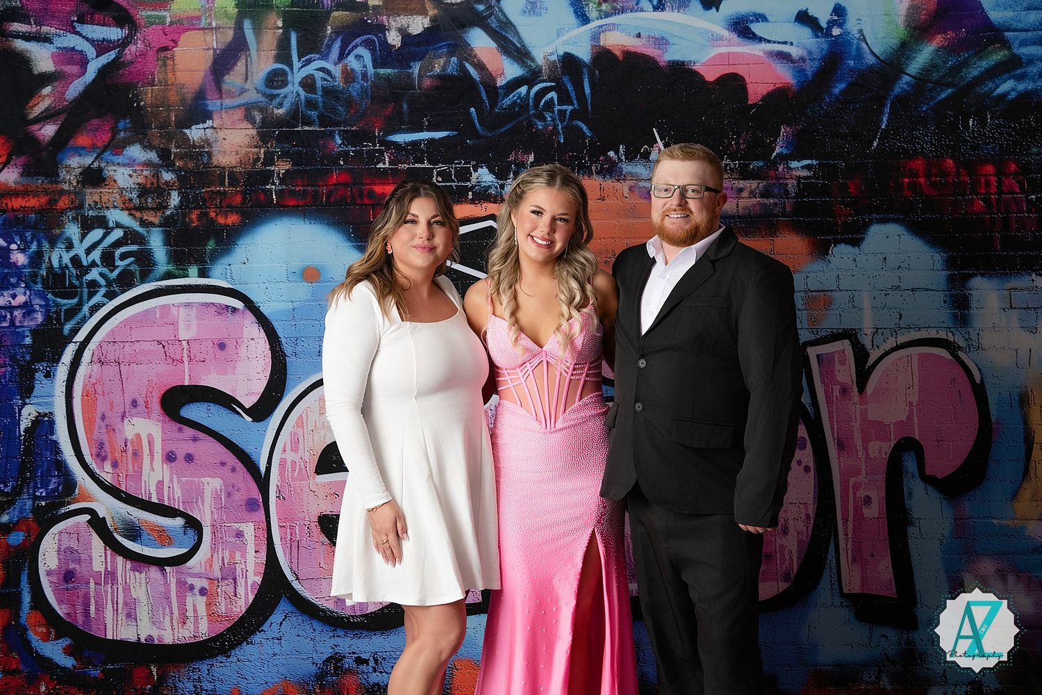 Three people pose together against a colorful graffiti wall backdrop with one wearing a pink formal dress.