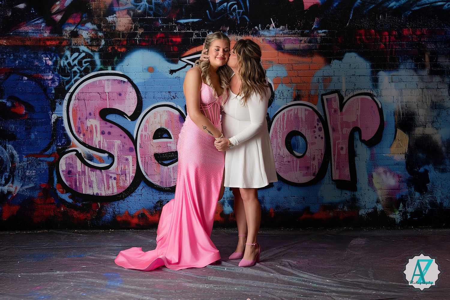 Two people in white and pink formal dresses pose together against a graffiti wall with vibrant pink spray paint designs.