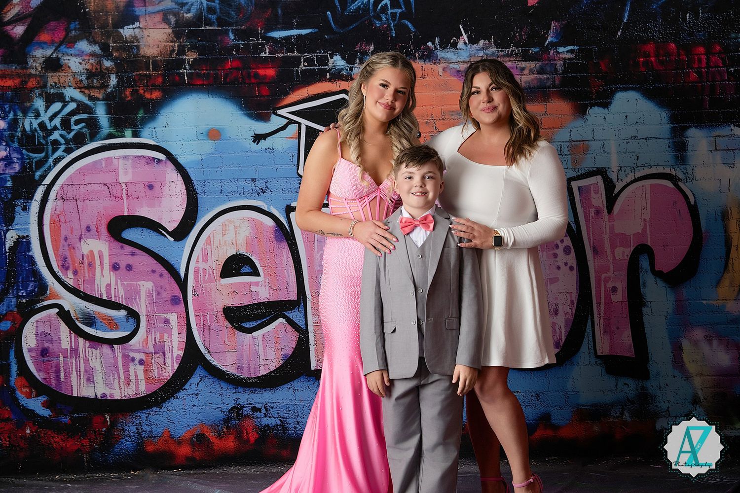 Three people pose in formal attire against a graffiti wall with vibrant pink street art.