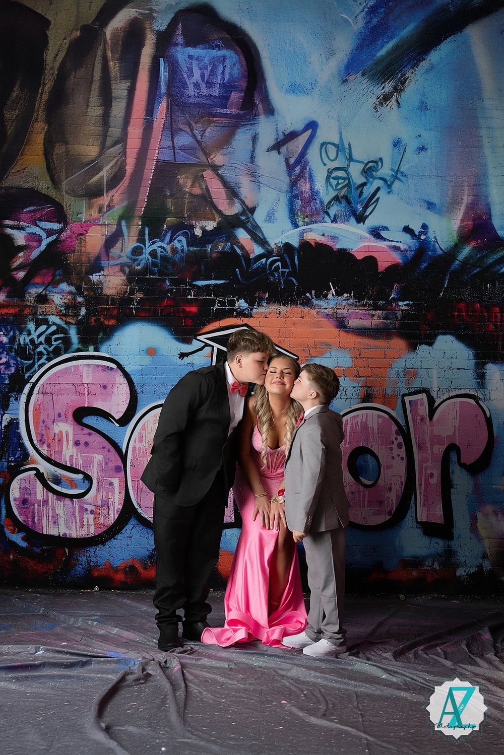 Three people pose together against a colorful graffiti wall with pink and blue urban street art in the background.