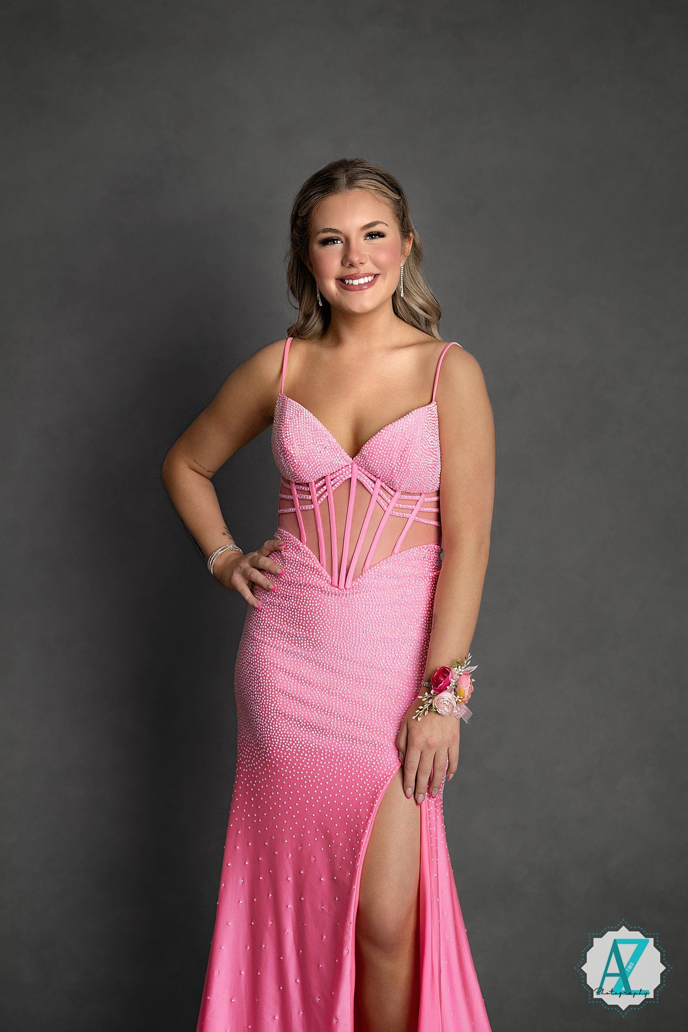 Young woman poses in a pink sparkly formal gown with corset bodice and side slit against grey backdrop.
