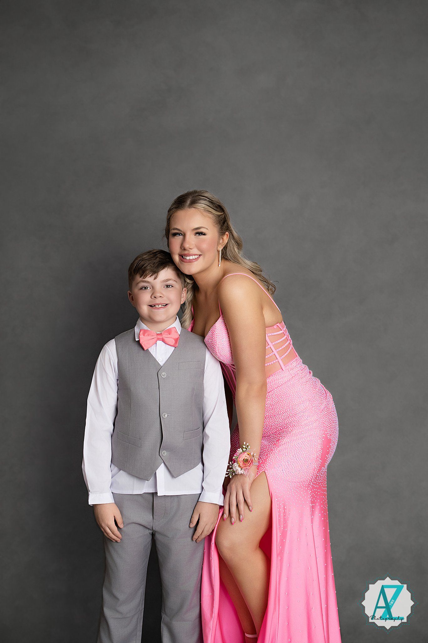 A young boy in formal gray attire poses with companion wearing a sparkly pink formal dress against a dark backdrop.