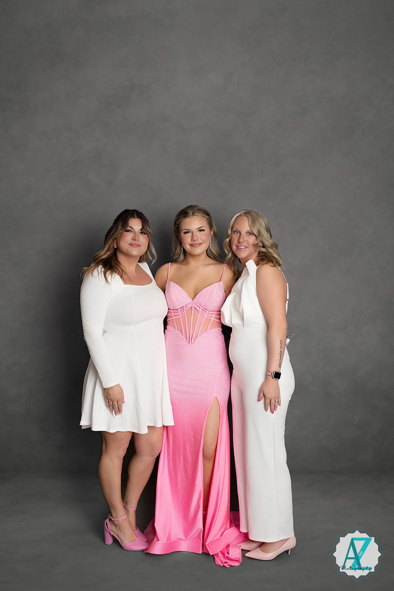 Three women pose together wearing elegant dresses - two in white and one in a pink gown with a leg slit against a gray backdrop.