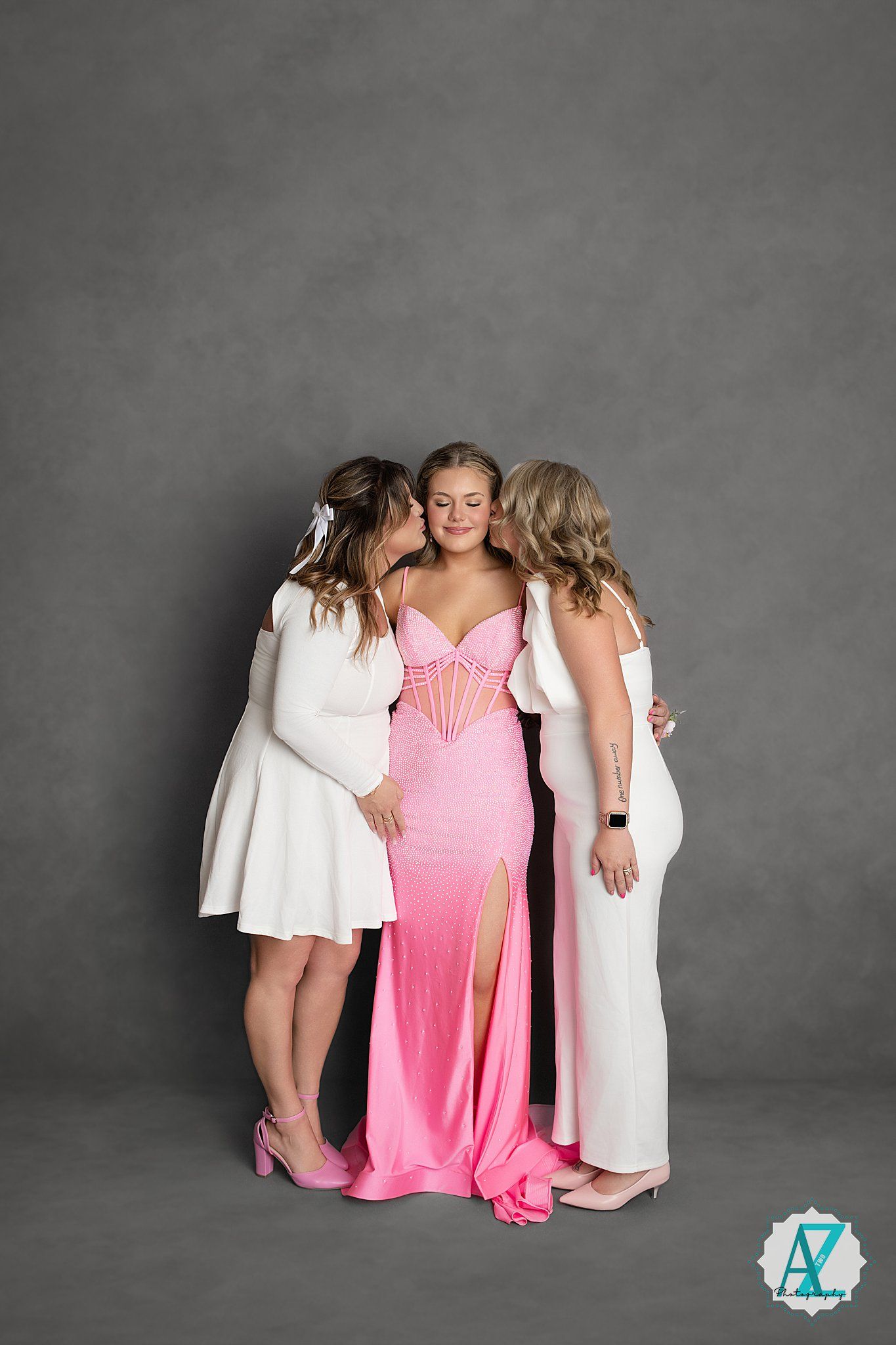 Three friends share a tender moment in front of a grey backdrop, with the center figure wearing a pink evening gown.