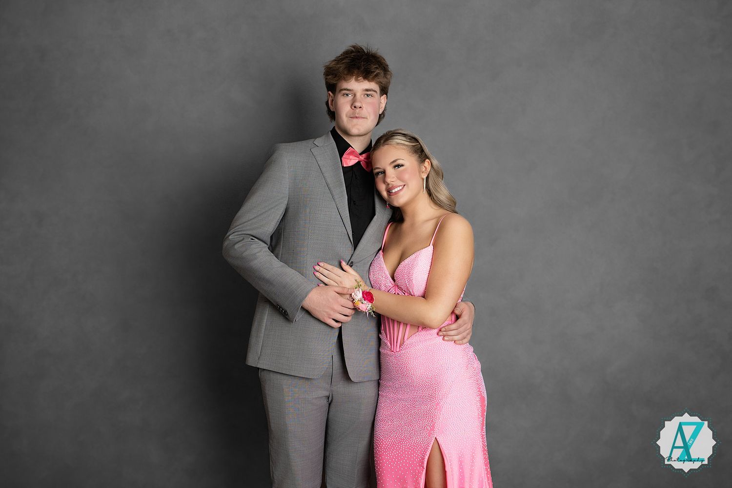 A couple poses together at a formal event, with one wearing a gray suit and the other in a pink gown with a slit.
