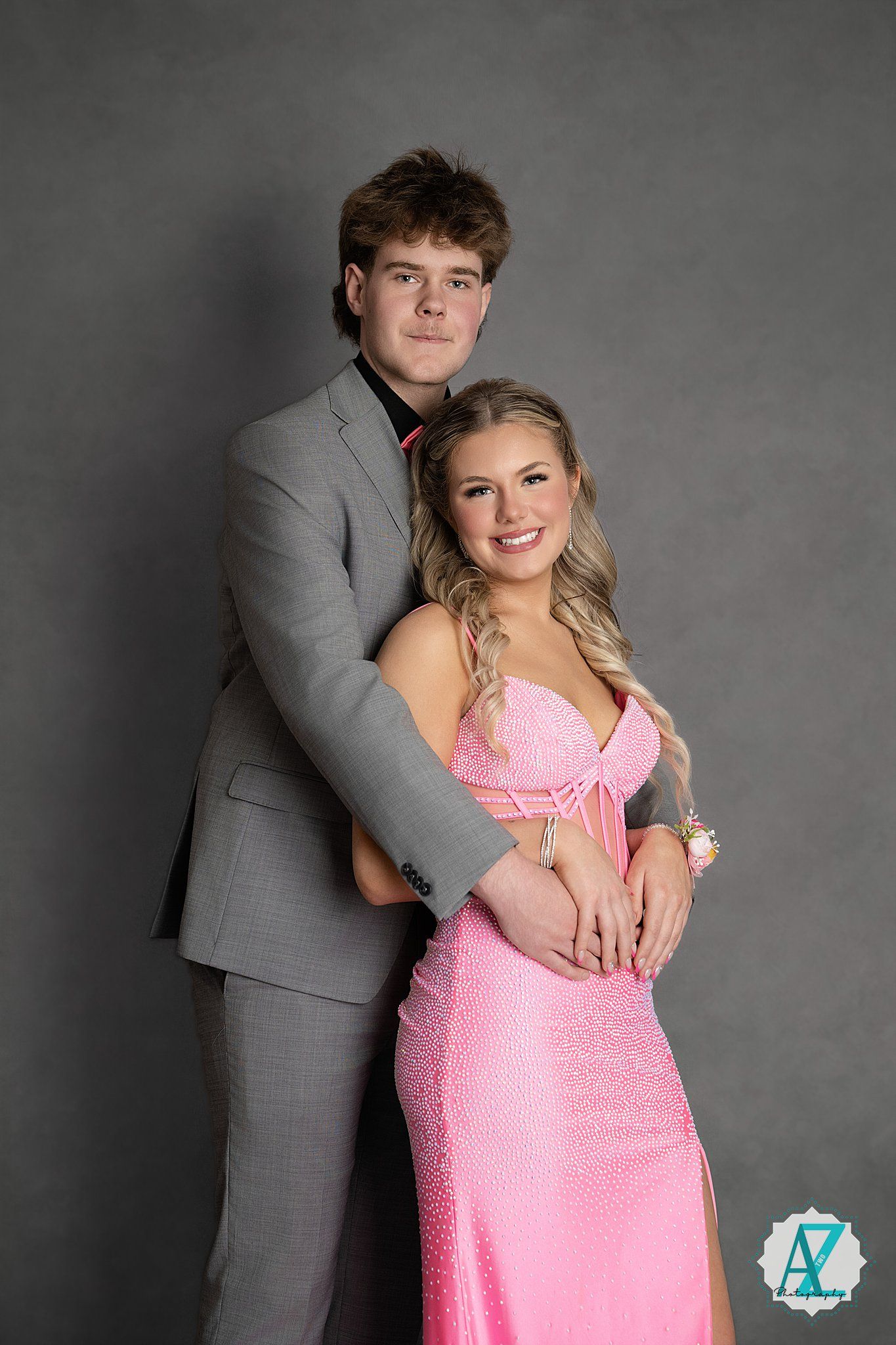 A couple poses together at a formal event, with her wearing a pink gown and him in a grey suit against a dark background.