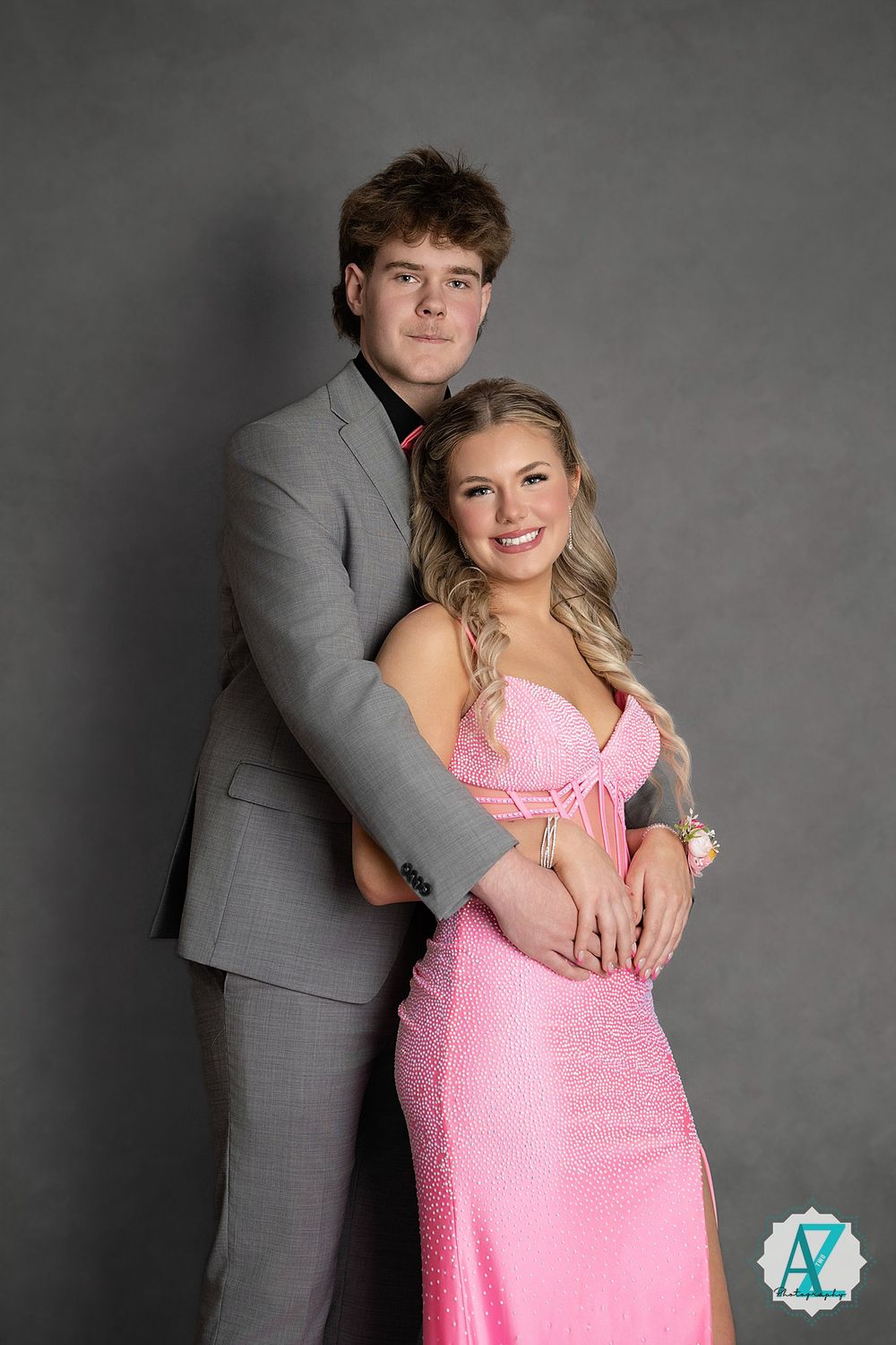A couple poses together at a formal event, with her wearing a pink gown and him in a grey suit against a dark background.