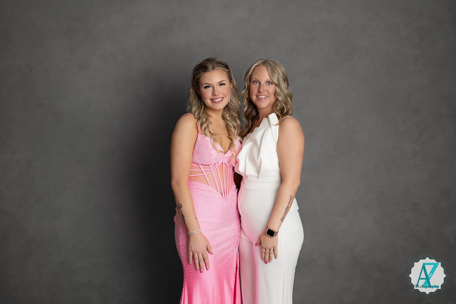 Two people in formal dresses pose together against a gray studio backdrop for an elegant portrait photo shoot.