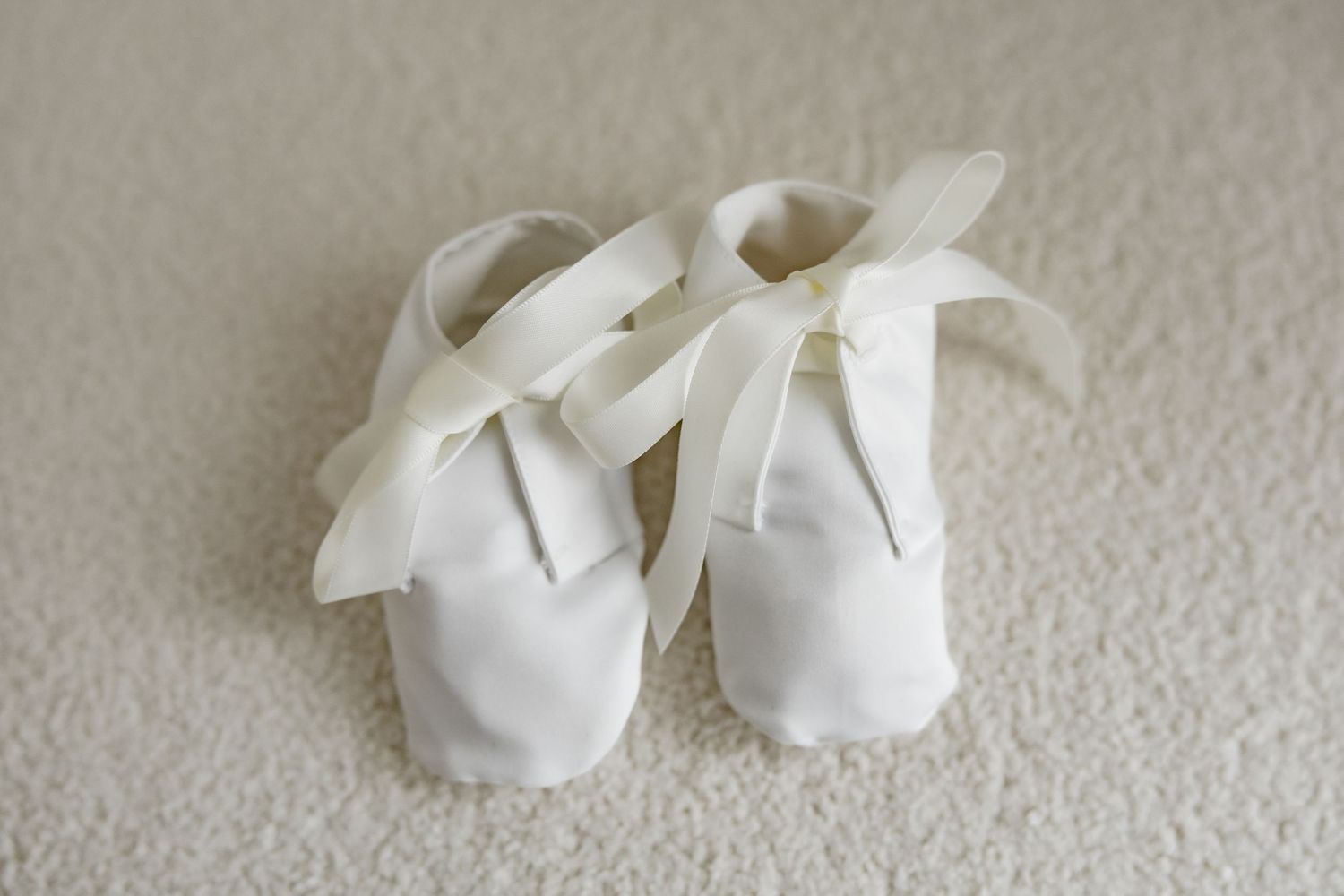 Small white baby baptism shoes with satin ribbon laces on light carpet background.