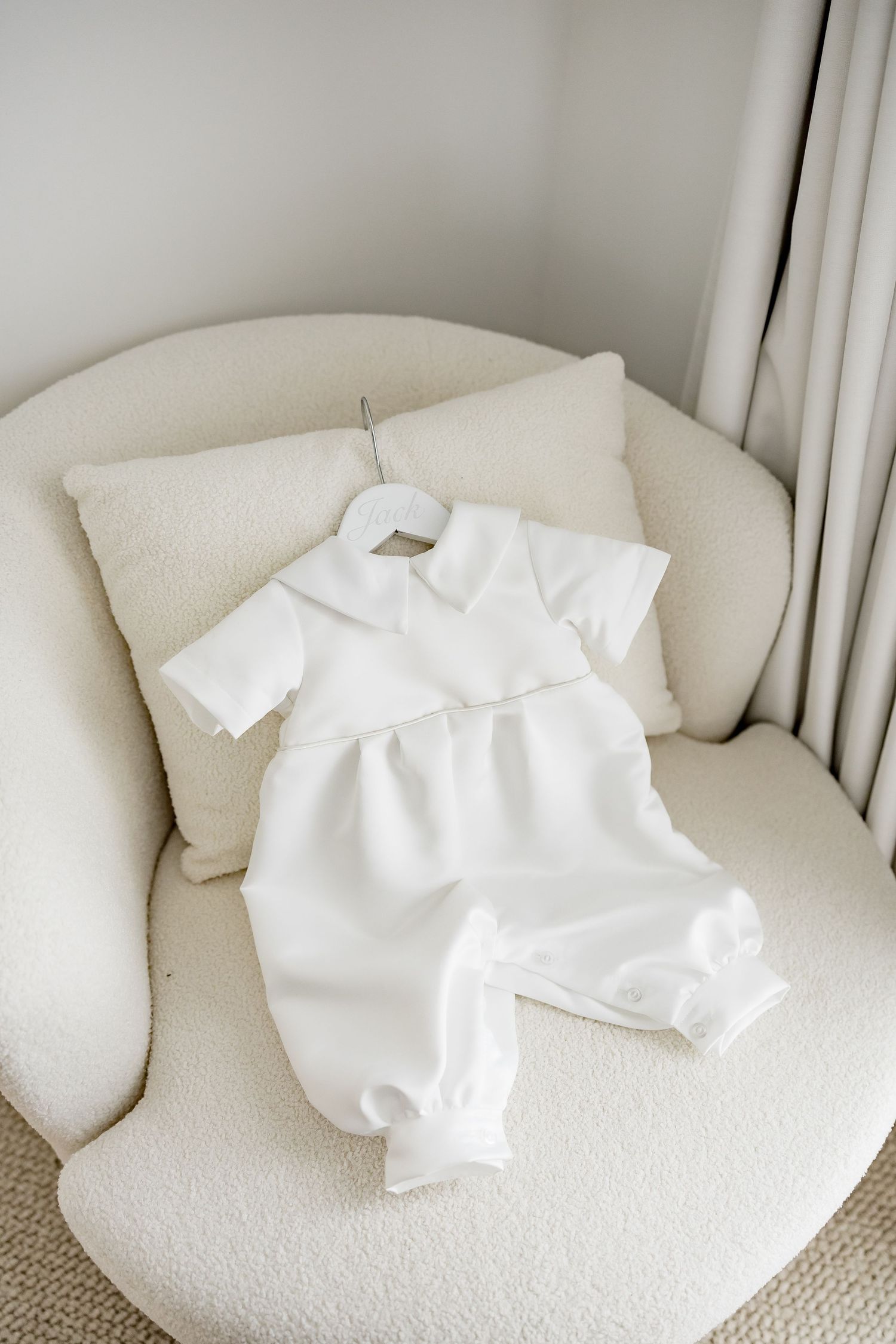A white baby christening gown or baptismal outfit laid out on a cream-colored armchair against neutral decor.