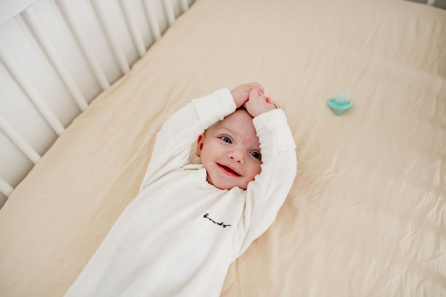 A baby wearing a white sleeper lying on beige bedding with arms raised and a teal pacifier nearby.