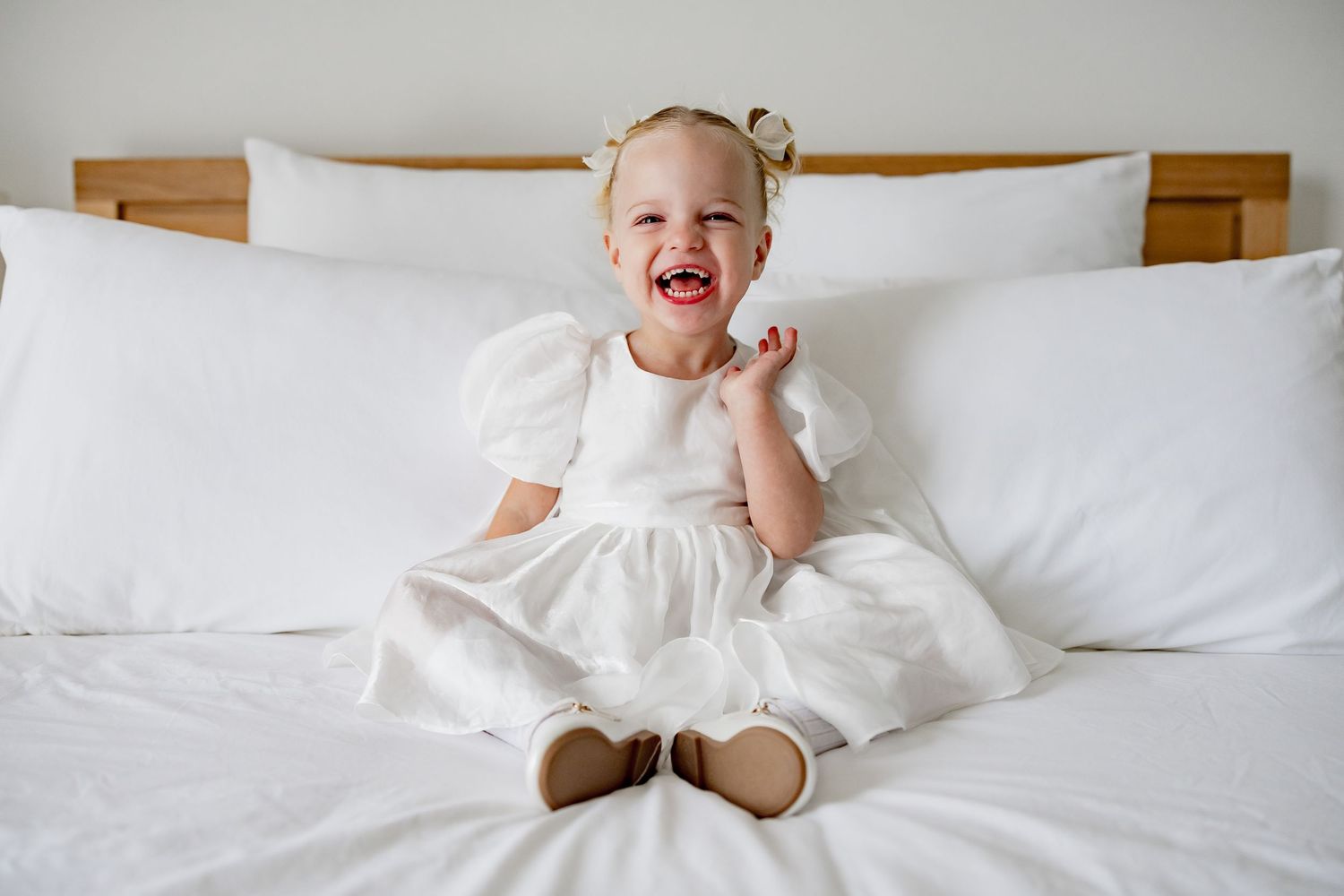A joyful child wearing a white dress sits on a bed with white pillows and linens, laughing with hand near face.