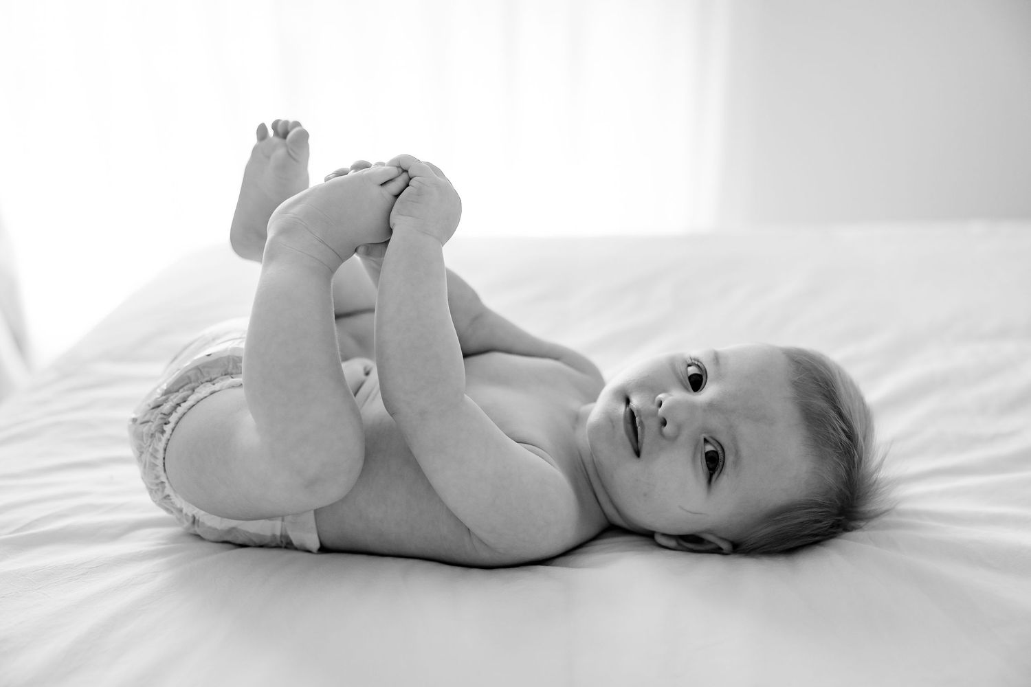 Black and white photo of an infant lying on white bedding while holding their feet with their hands and smiling.