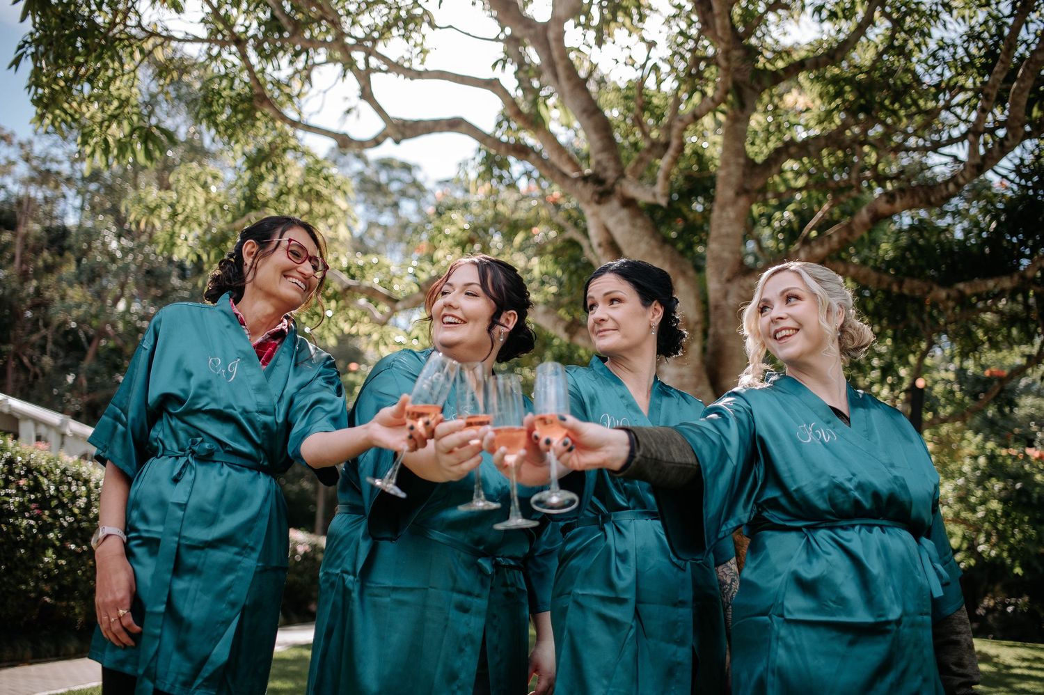 Bridesmaids in matching teal robes laughing and toasting with wine glasses in an outdoor garden setting.