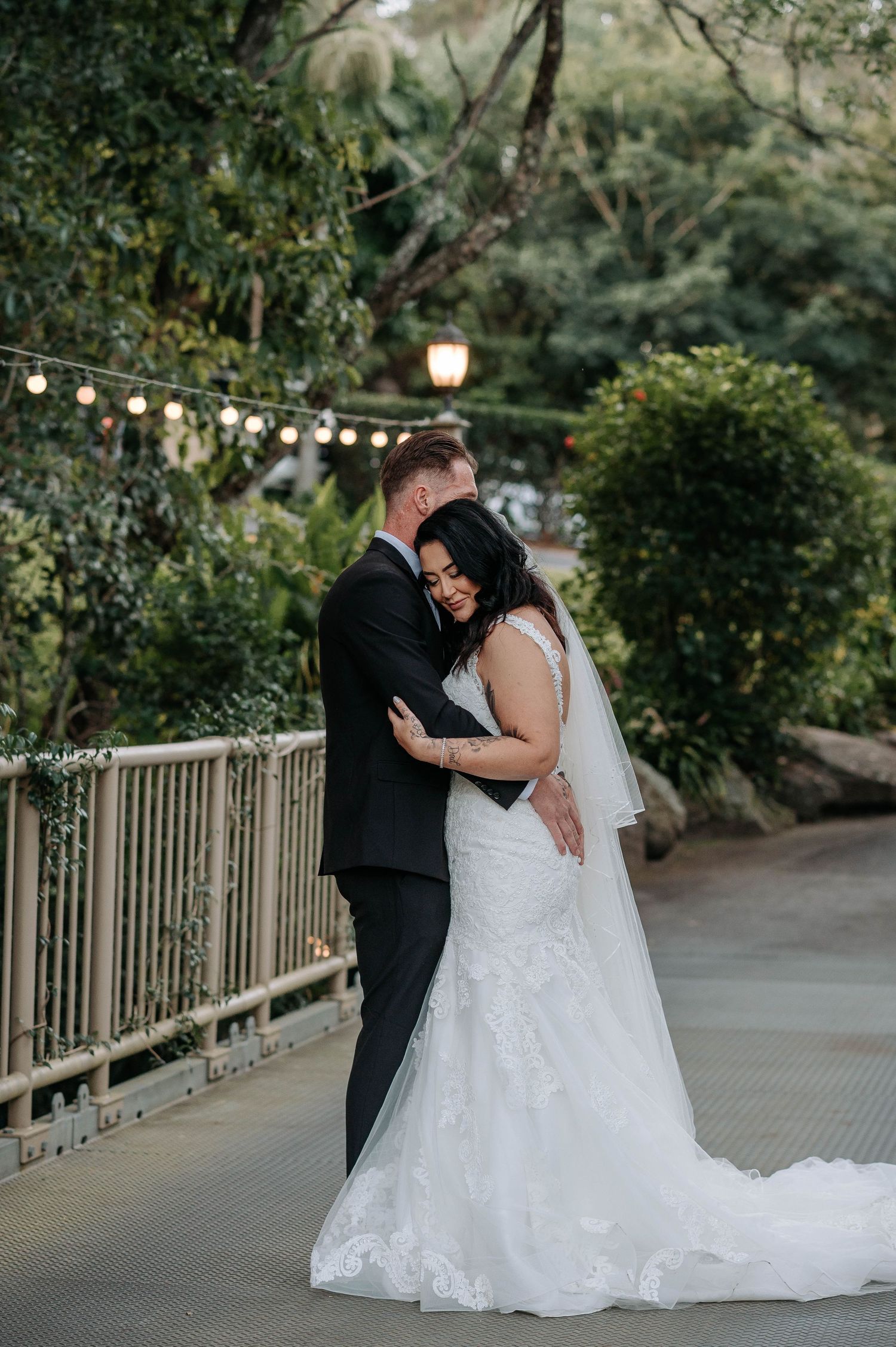 Romantic couple embraces on a garden path under string lights and greenery during sunset wedding photoshoot.