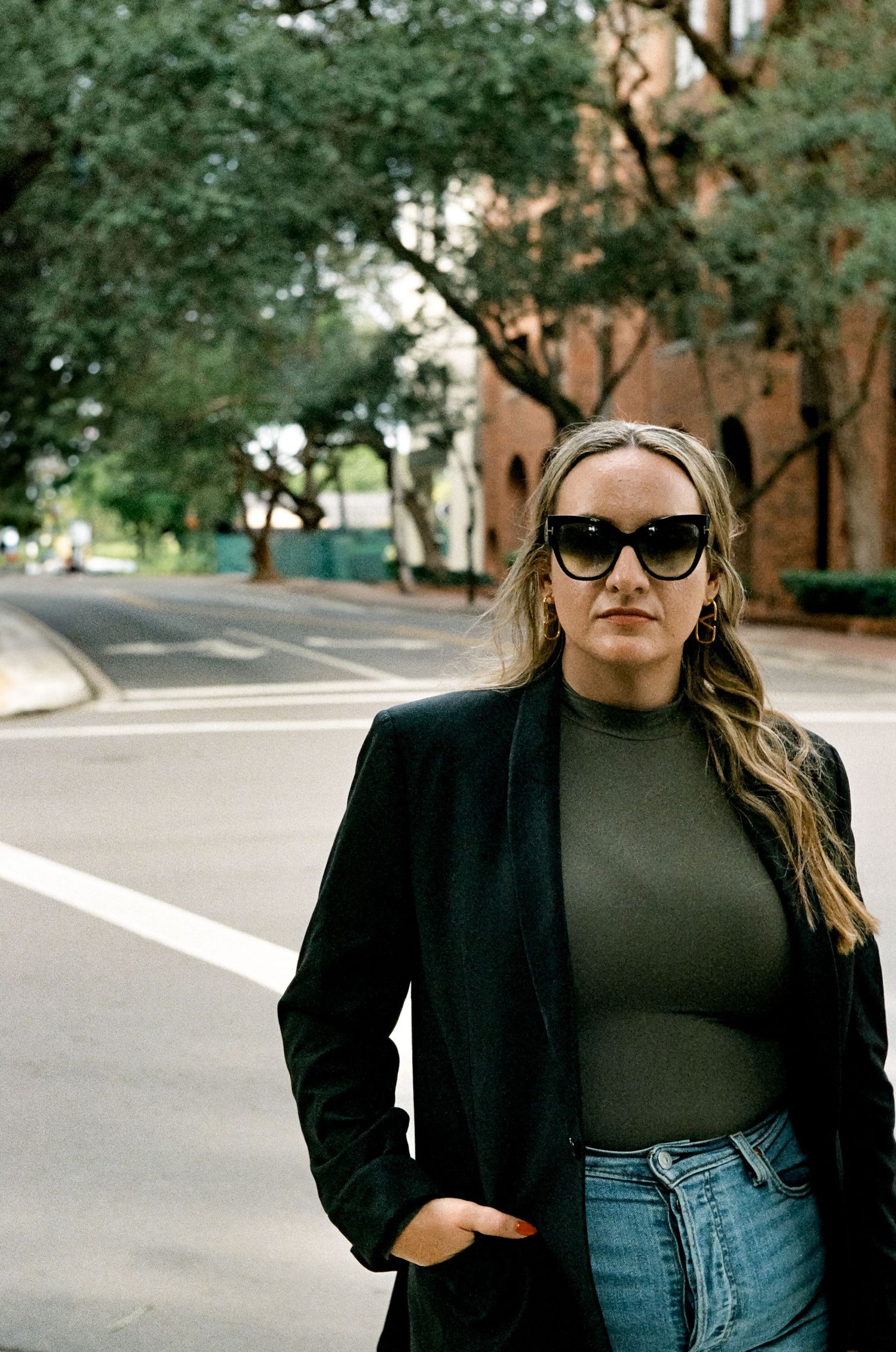A person wearing sunglasses, black blazer and olive top stands on a tree-lined street corner in an urban setting.