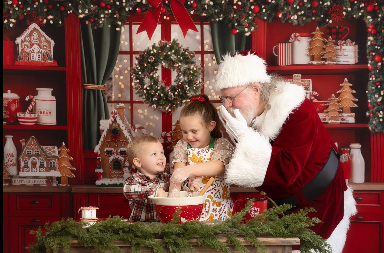 Two children sit with Santa Claus in a festive Christmas kitchen decorated with holiday ornaments and garland.