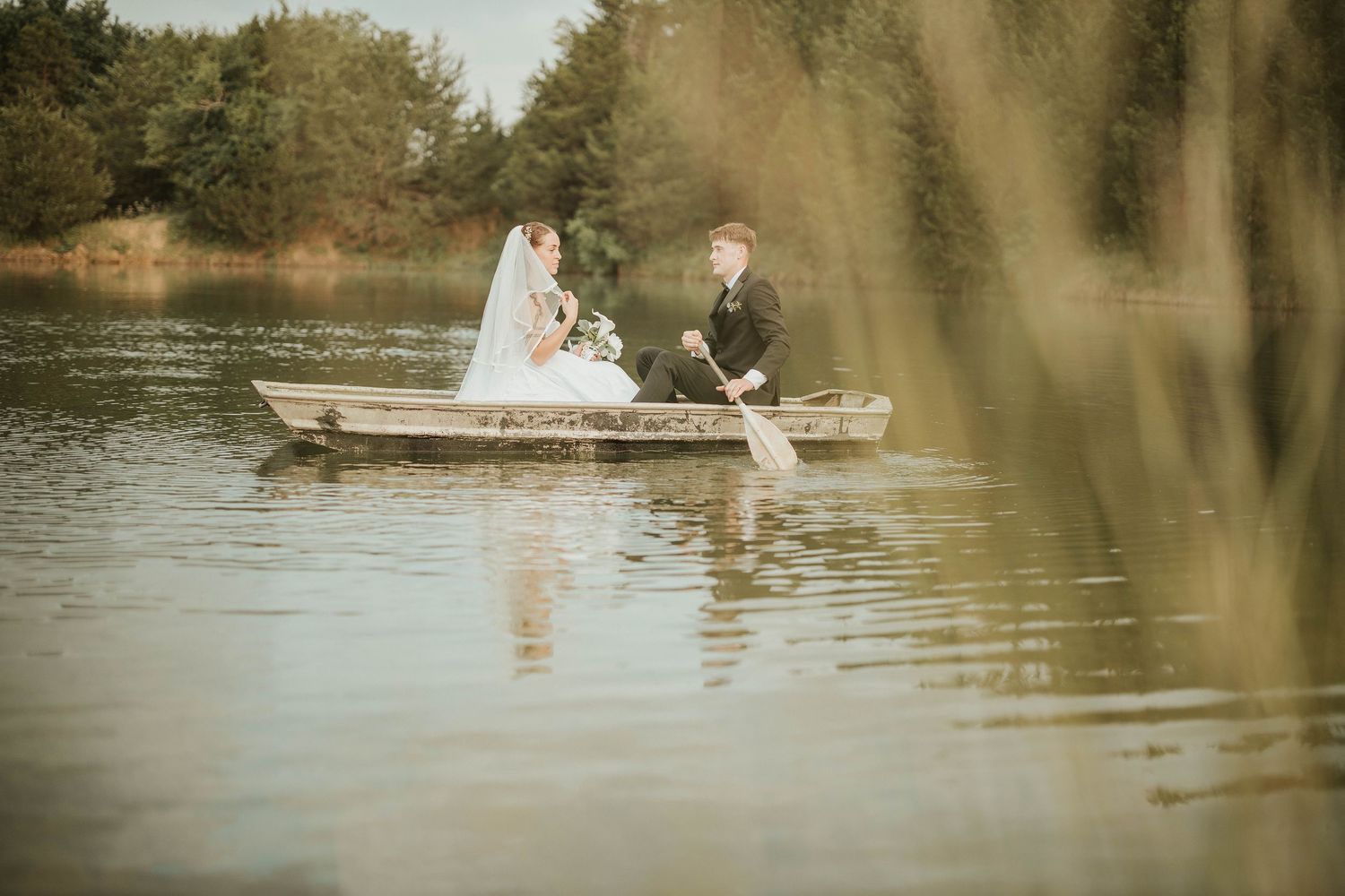 A wedding couple sits in a wooden rowboat on a tranquil lake surrounded by nature.