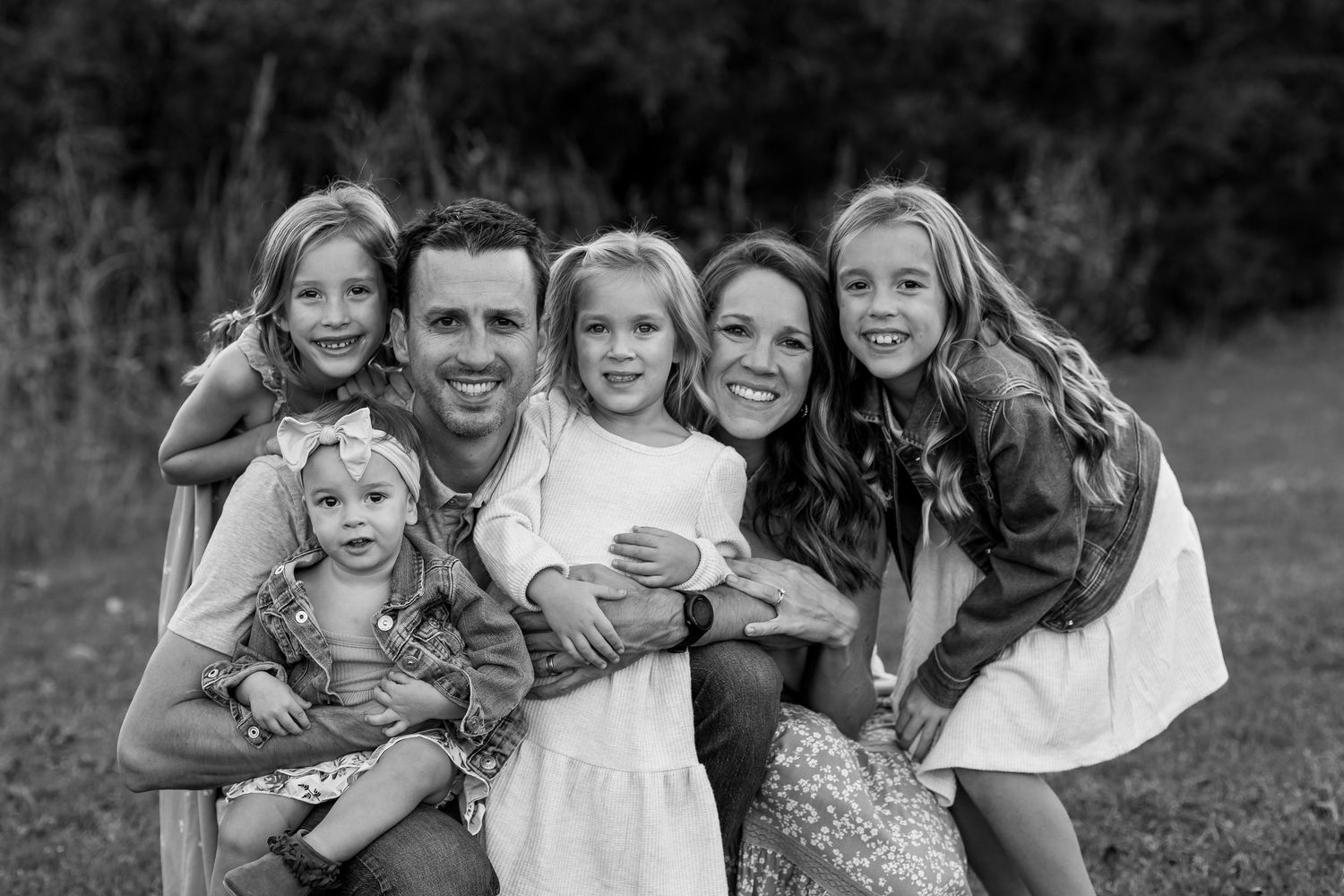 A black and white outdoor portrait of a close-knit family group hugging and smiling together in nature.