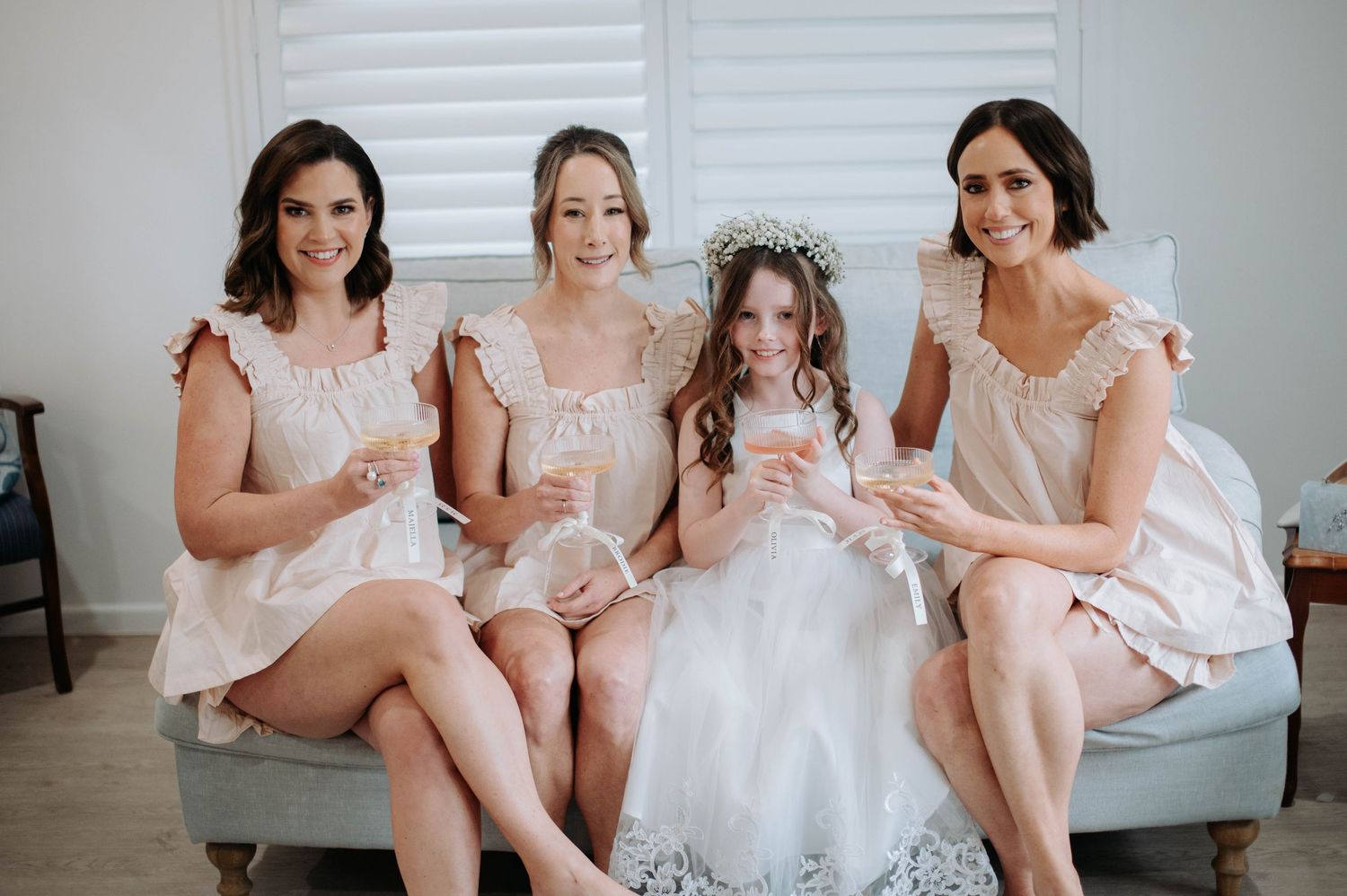 Wedding party members in blush pink dresses sit together on gray couch during bridal preparations.