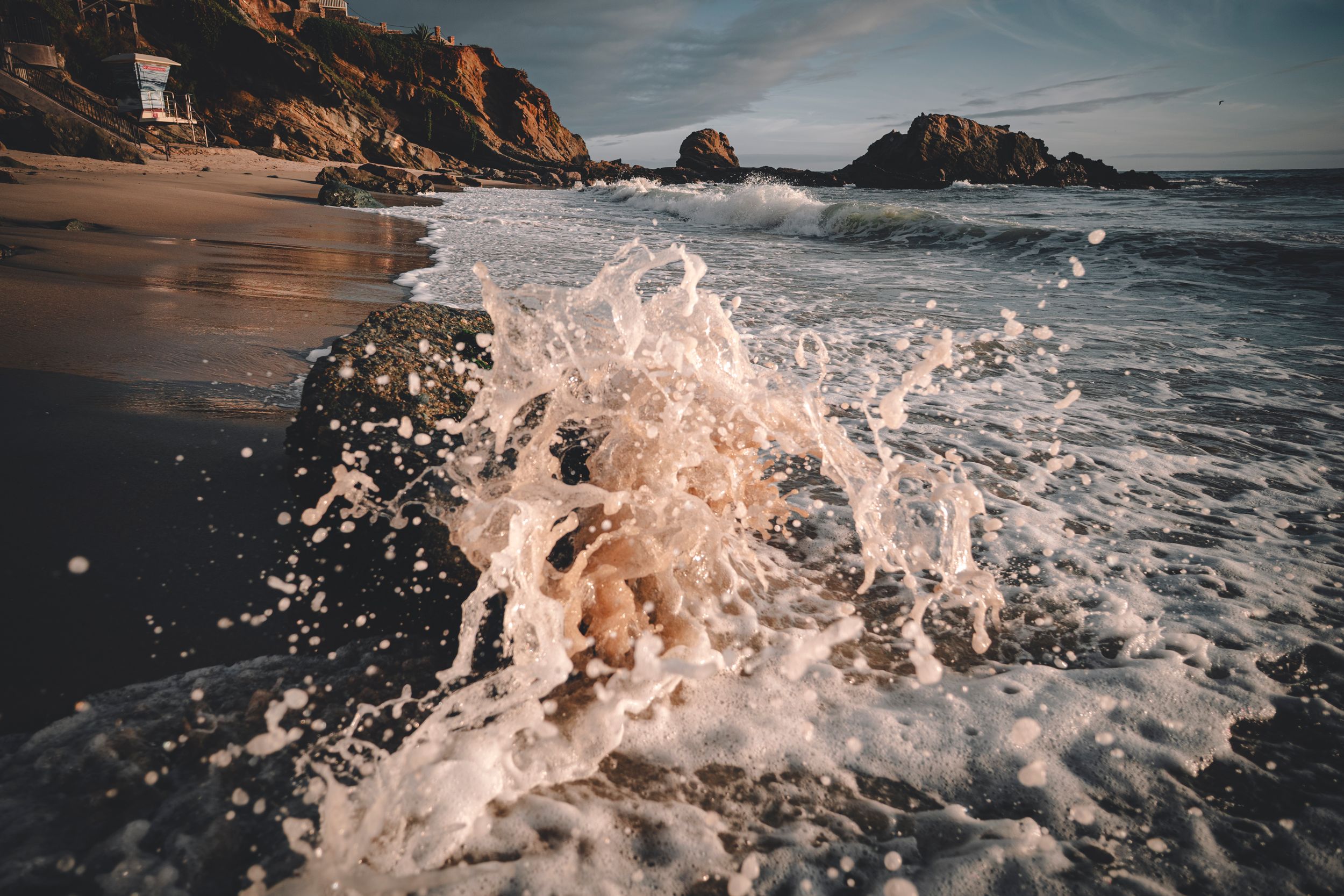 Ocean wave splashing dramatically on a rocky California beach with golden cliffs in the background.