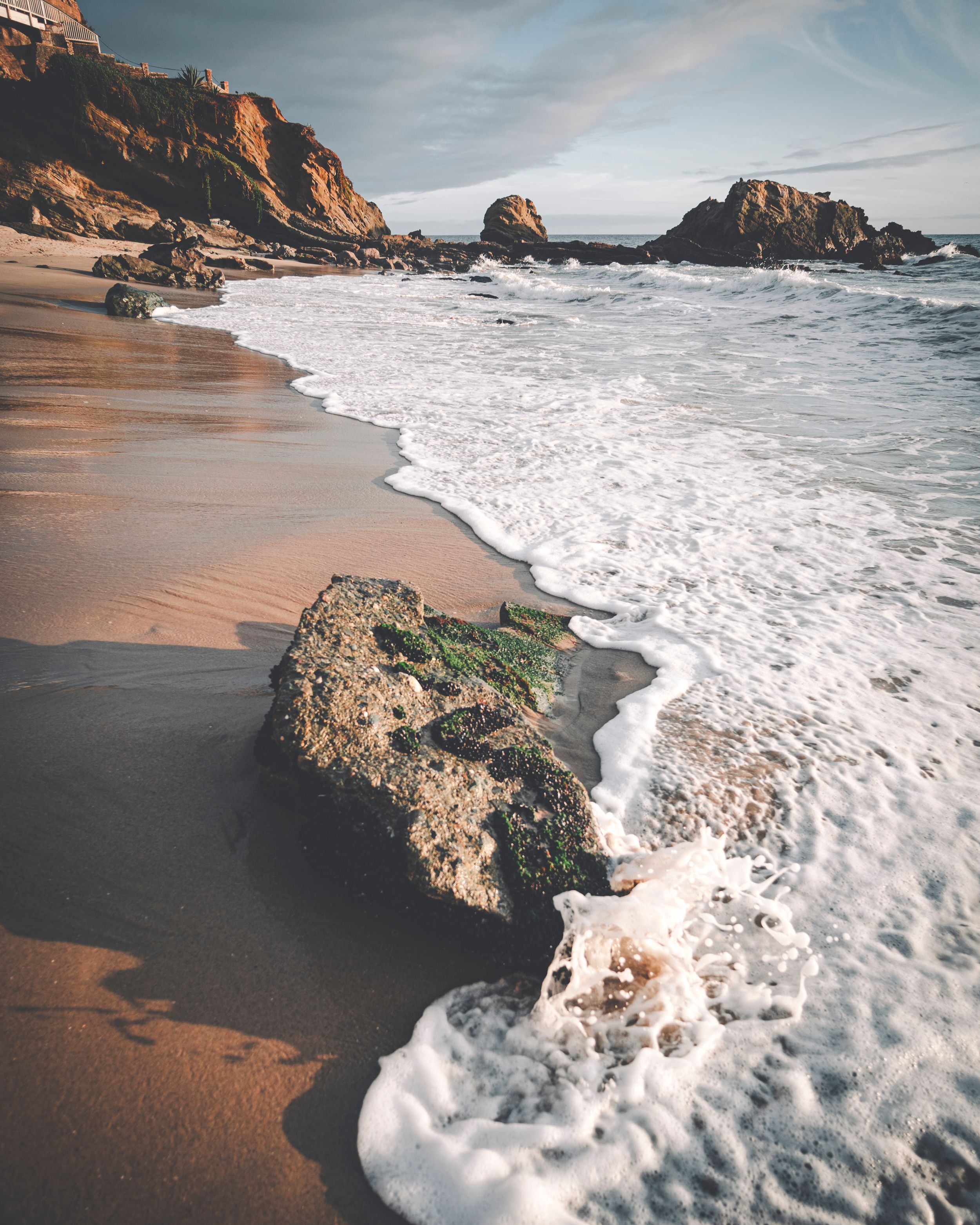 Foamy ocean waves washing over rocks on a sandy California beach with rugged cliffs.