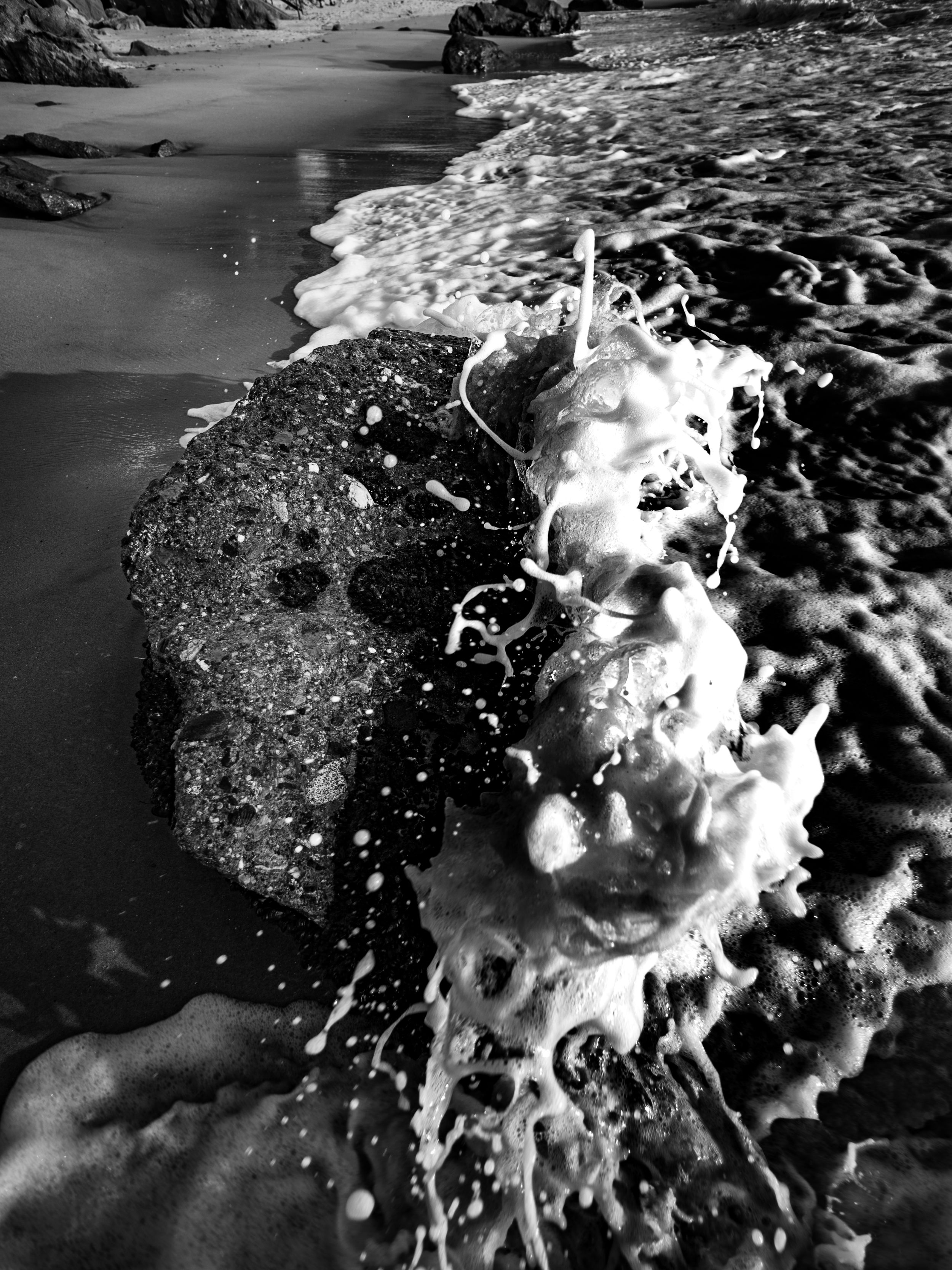 Black and white ocean wave splashing dramatically over a large rock on a sandy beach.