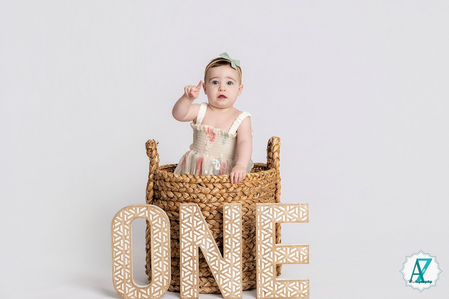 Baby sitting in woven basket with gold glitter ONE letters for first birthday photoshoot against white backdrop.