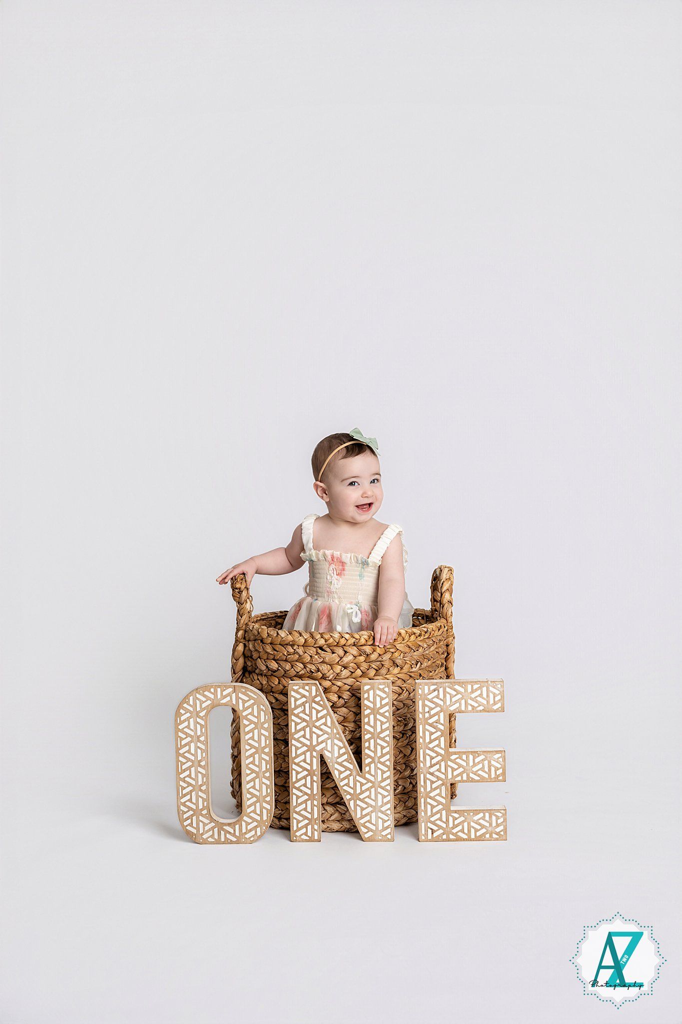 Baby in light outfit sitting in woven basket with gold sparkly ONE letters against white background.