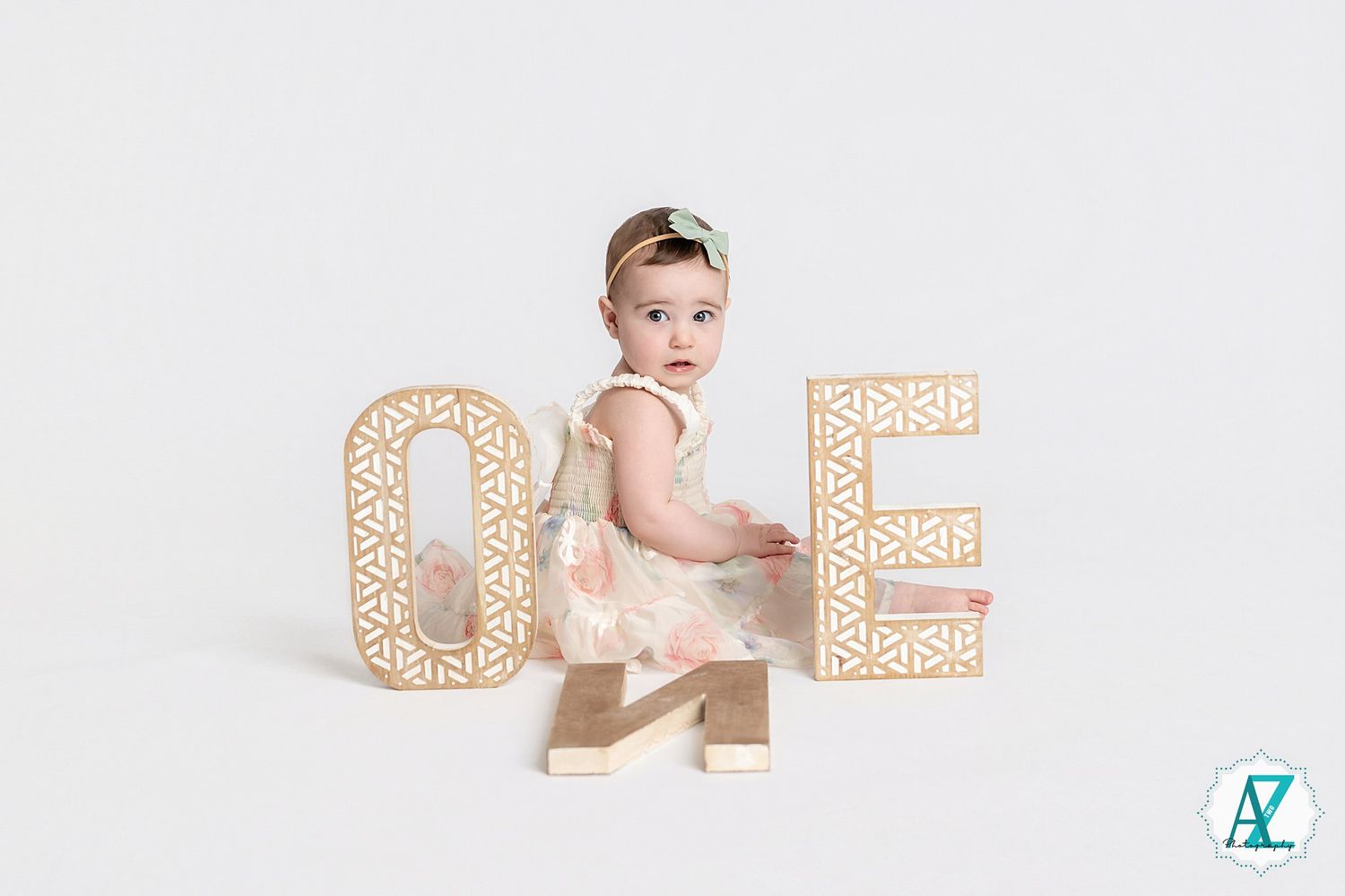 Baby in light dress poses beside decorative gold number one letters against white background.