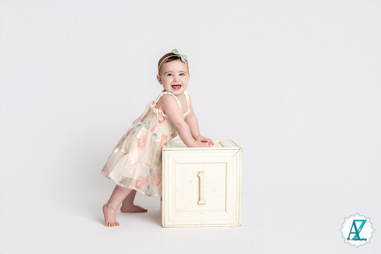 Baby in cream dress standing next to white number one block against plain background.