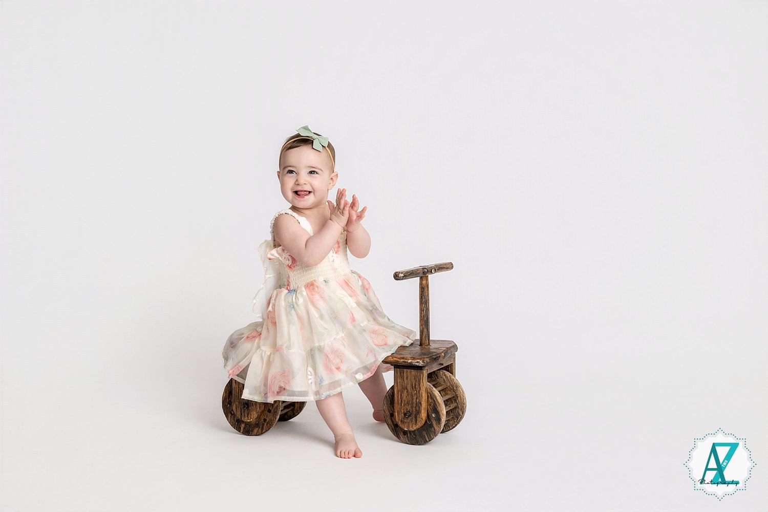 Baby wearing cream dress posing on vintage wooden tricycle against white background.