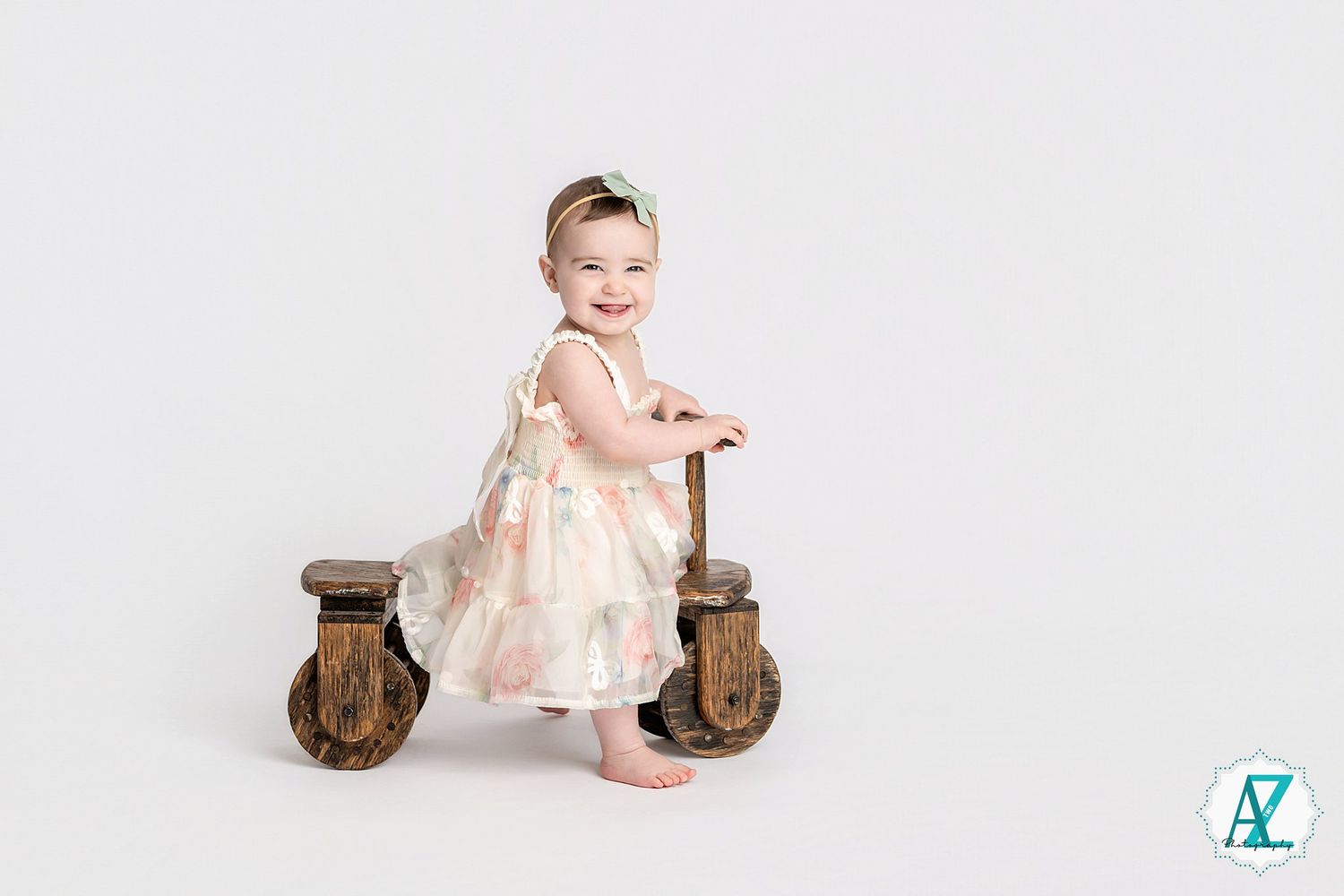 Small child in floral dress sits on wooden stool against plain white backdrop for portrait.