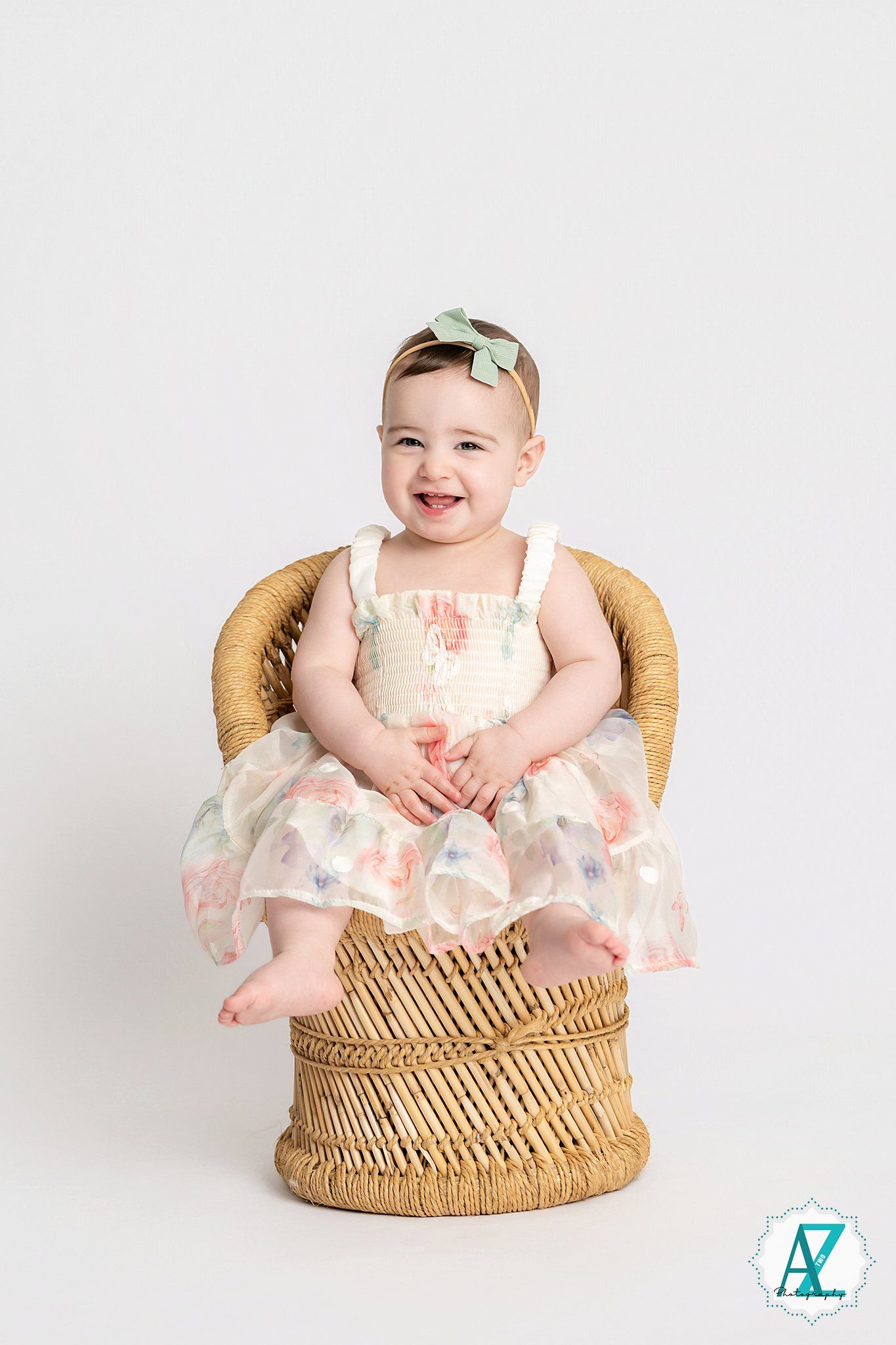 Baby seated in a round wicker basket wearing a delicate floral dress against a white backdrop.