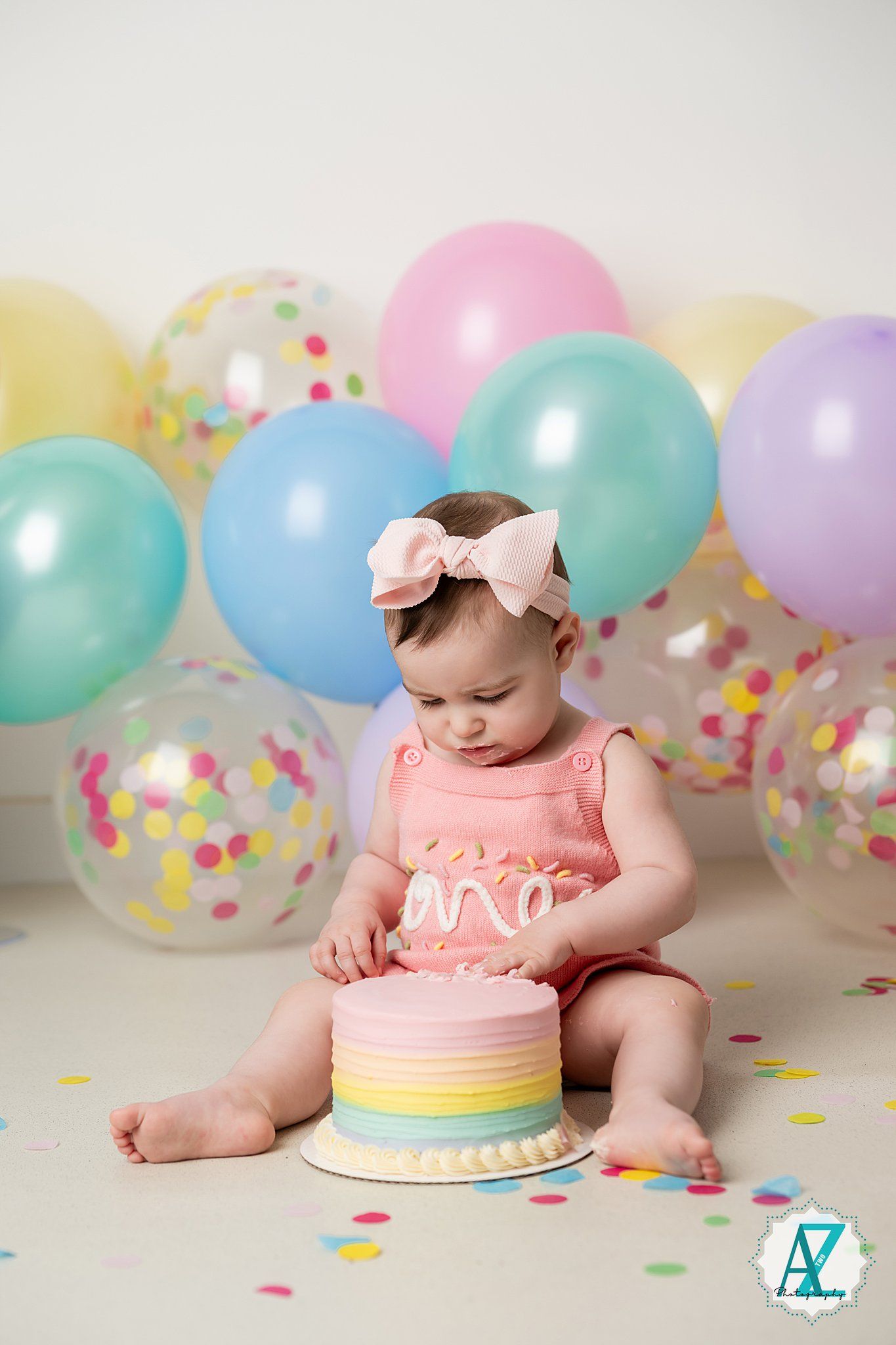 Baby in pink outfit with bow at first birthday cake smash session with pastel balloons.